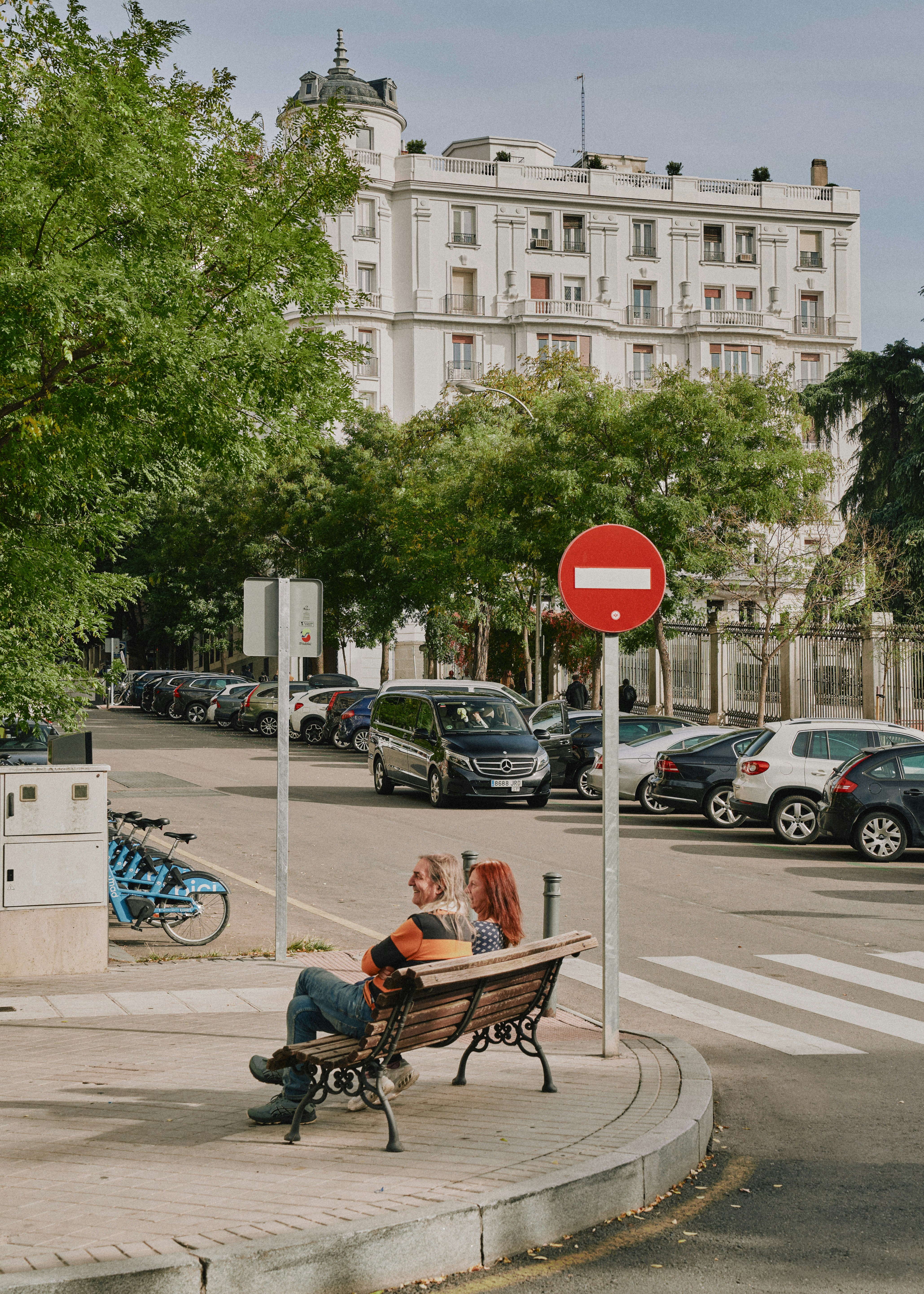 Couple sitting on park bench with cars and building.