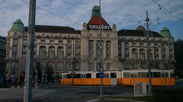 The Art Nouveau facade of the Gellert building, its name spelled out above the entrance, with a yellow tram passing in front.