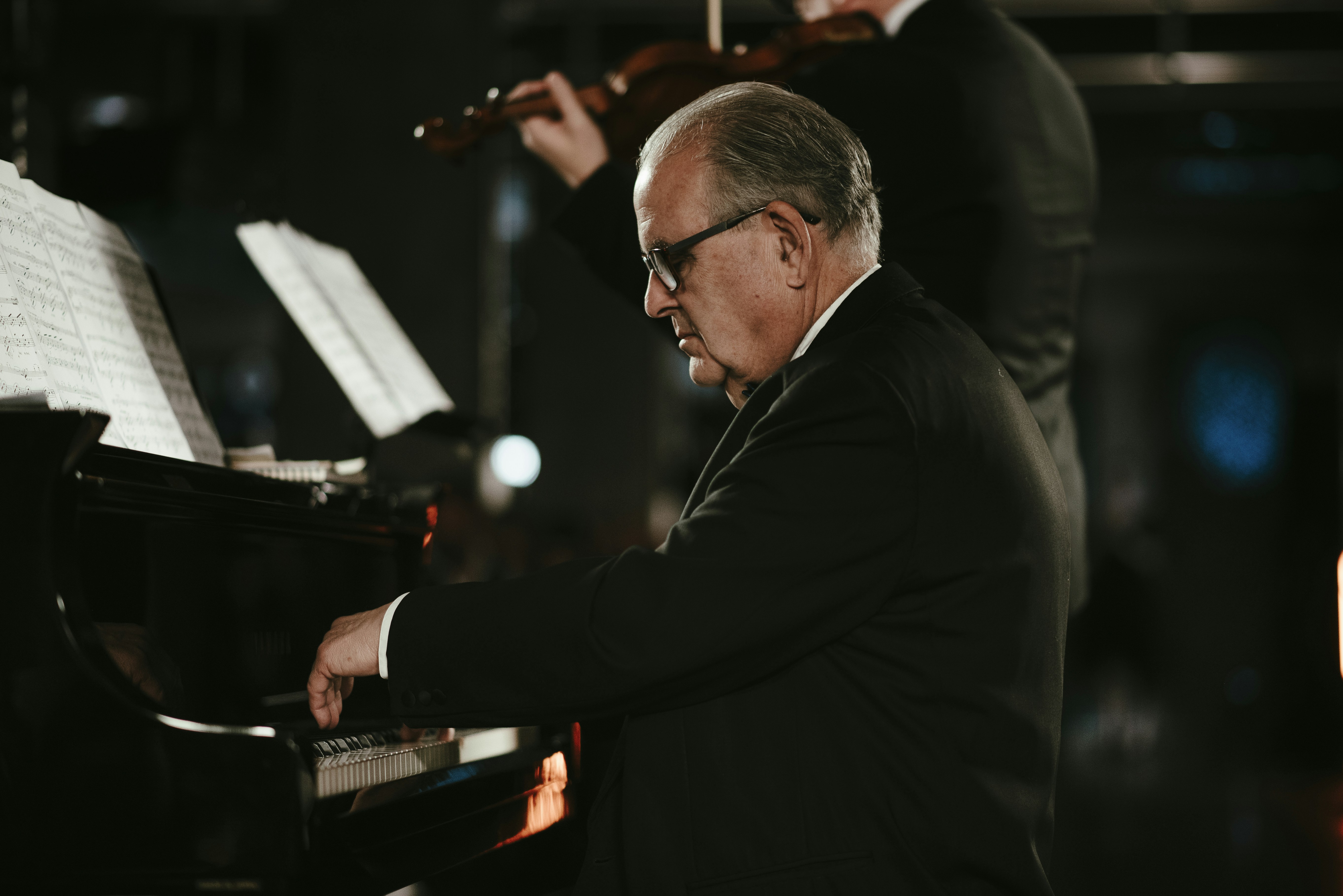 Pianist spielt Flügel mit einem Geiger im Hintergrund.