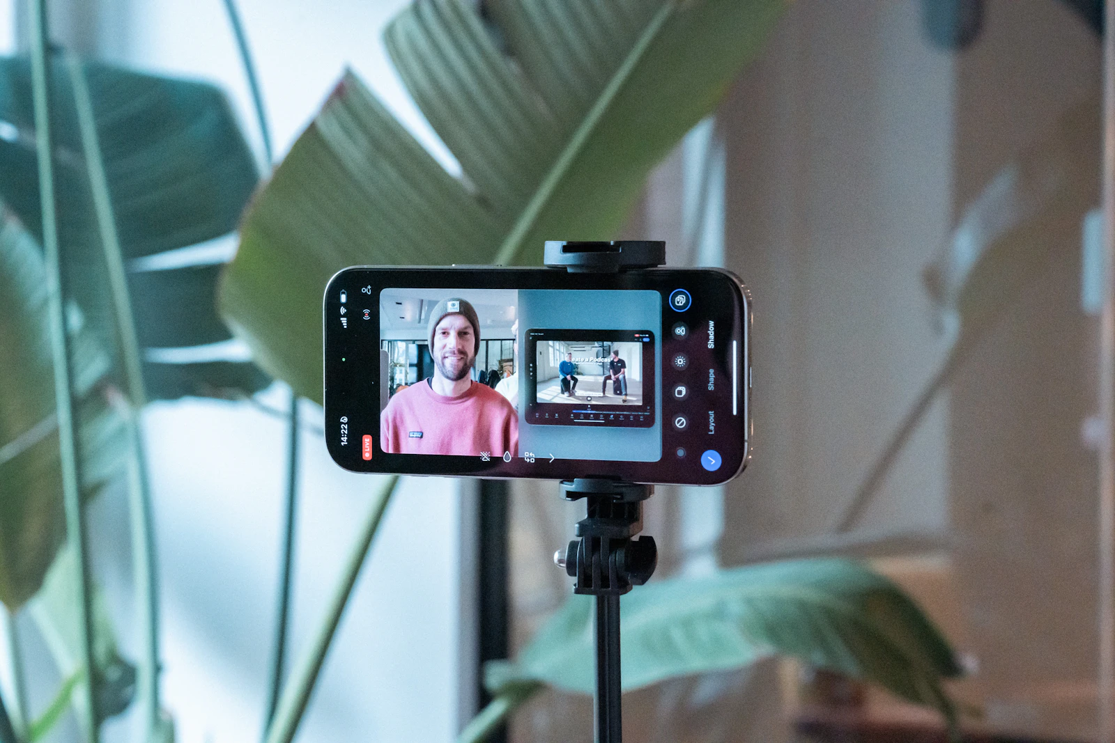 A smartphone on a tripod recording a man in a pink shirt in a bright room with plants