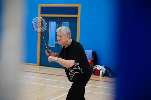 Elderly woman playing badminton in a gymnasium.