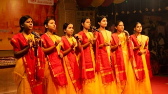 Six young women in yellow dresses and red sashes singing.