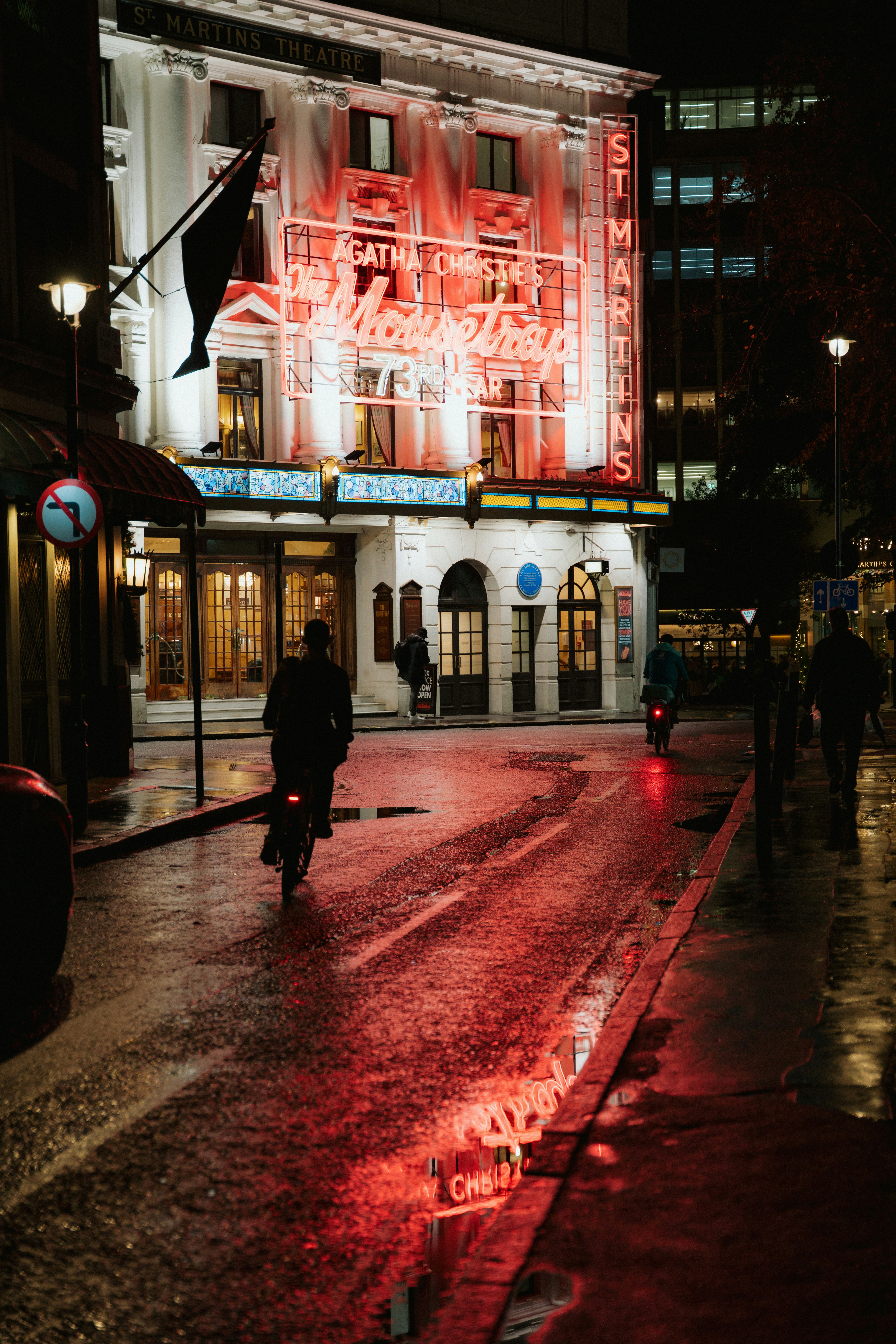 Wet street reflects neon lights outside theatre at night.