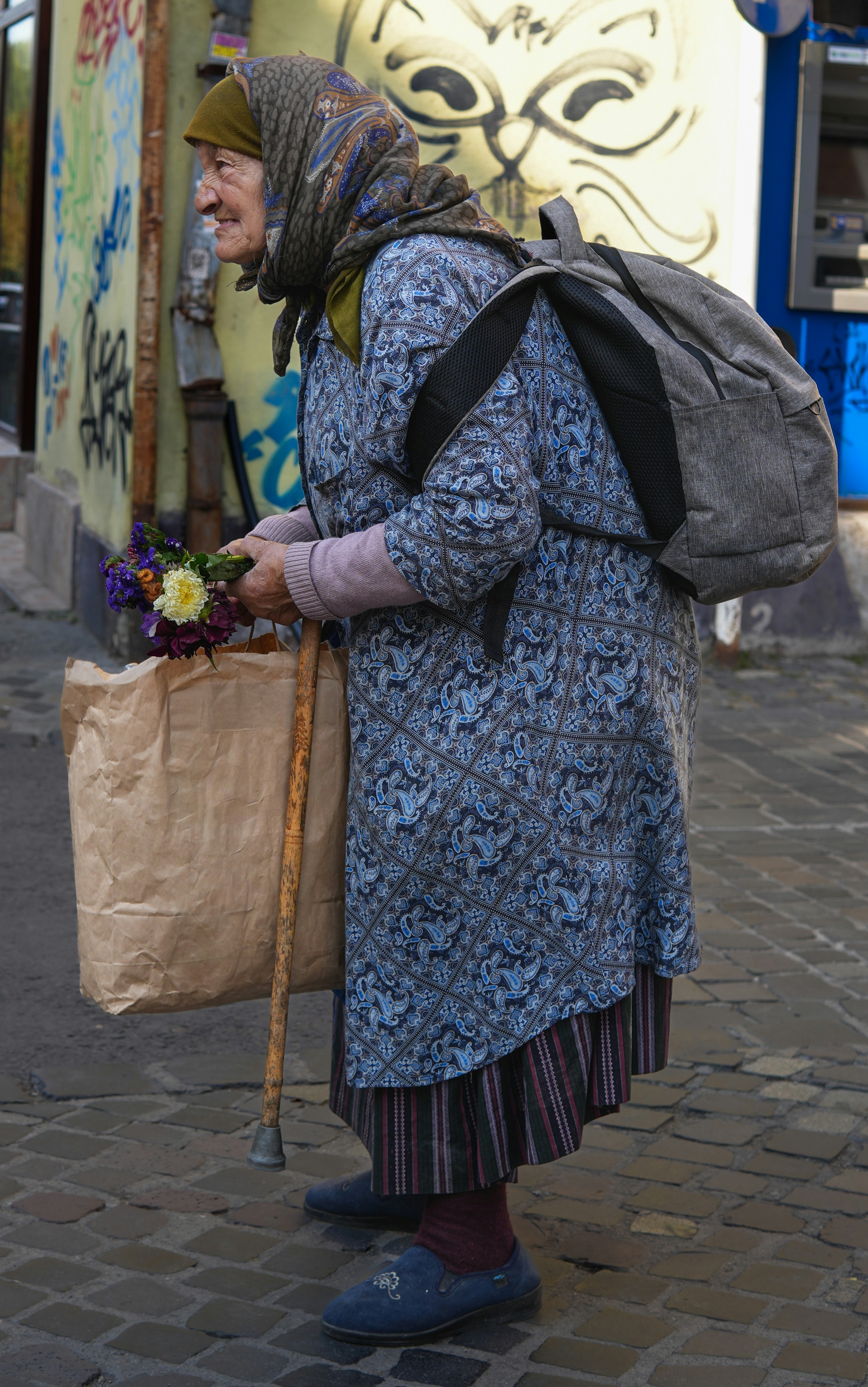 Elderly woman with a walking stick, flowers, and backpack.