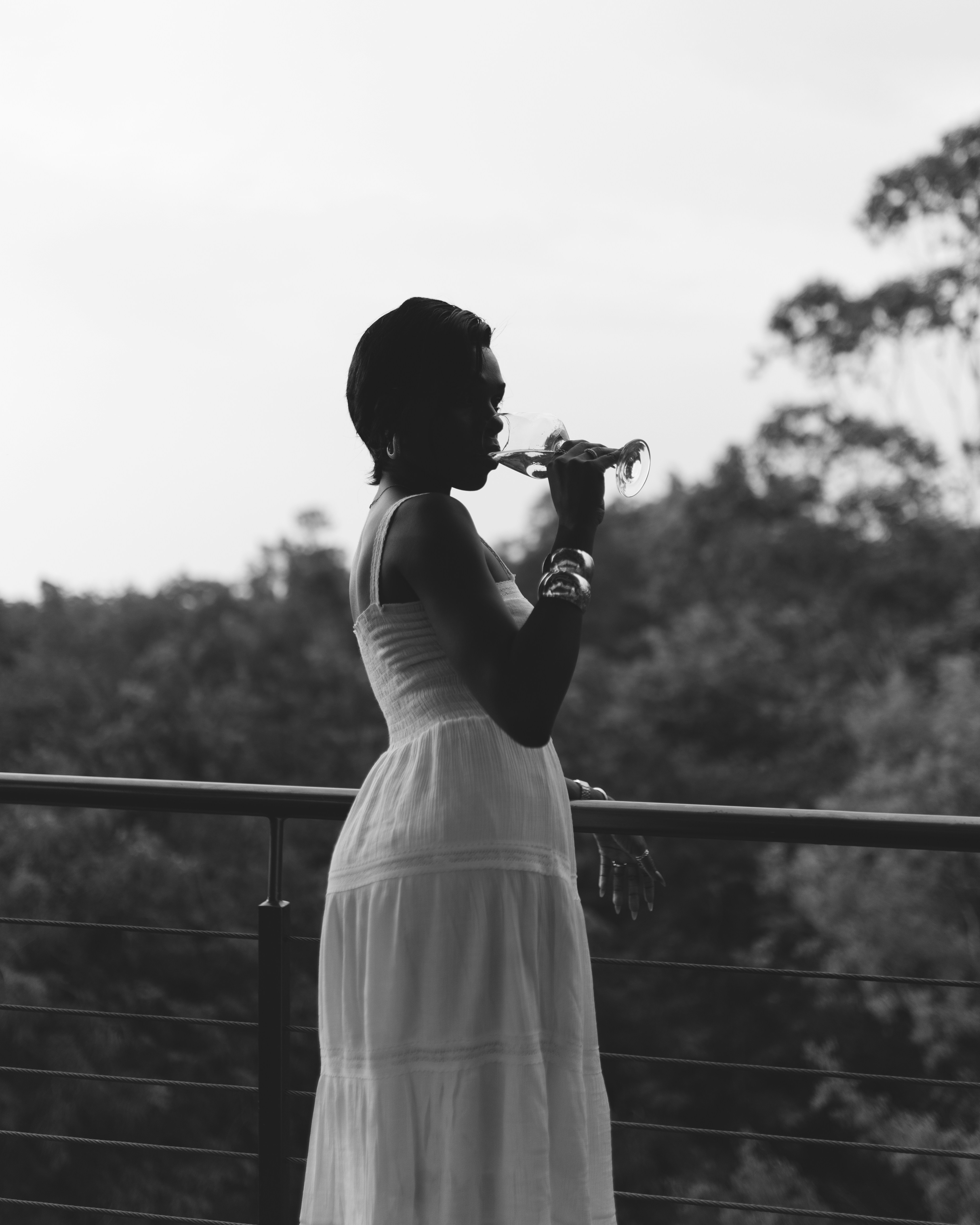 Woman in white dress drinks from glass on balcony.