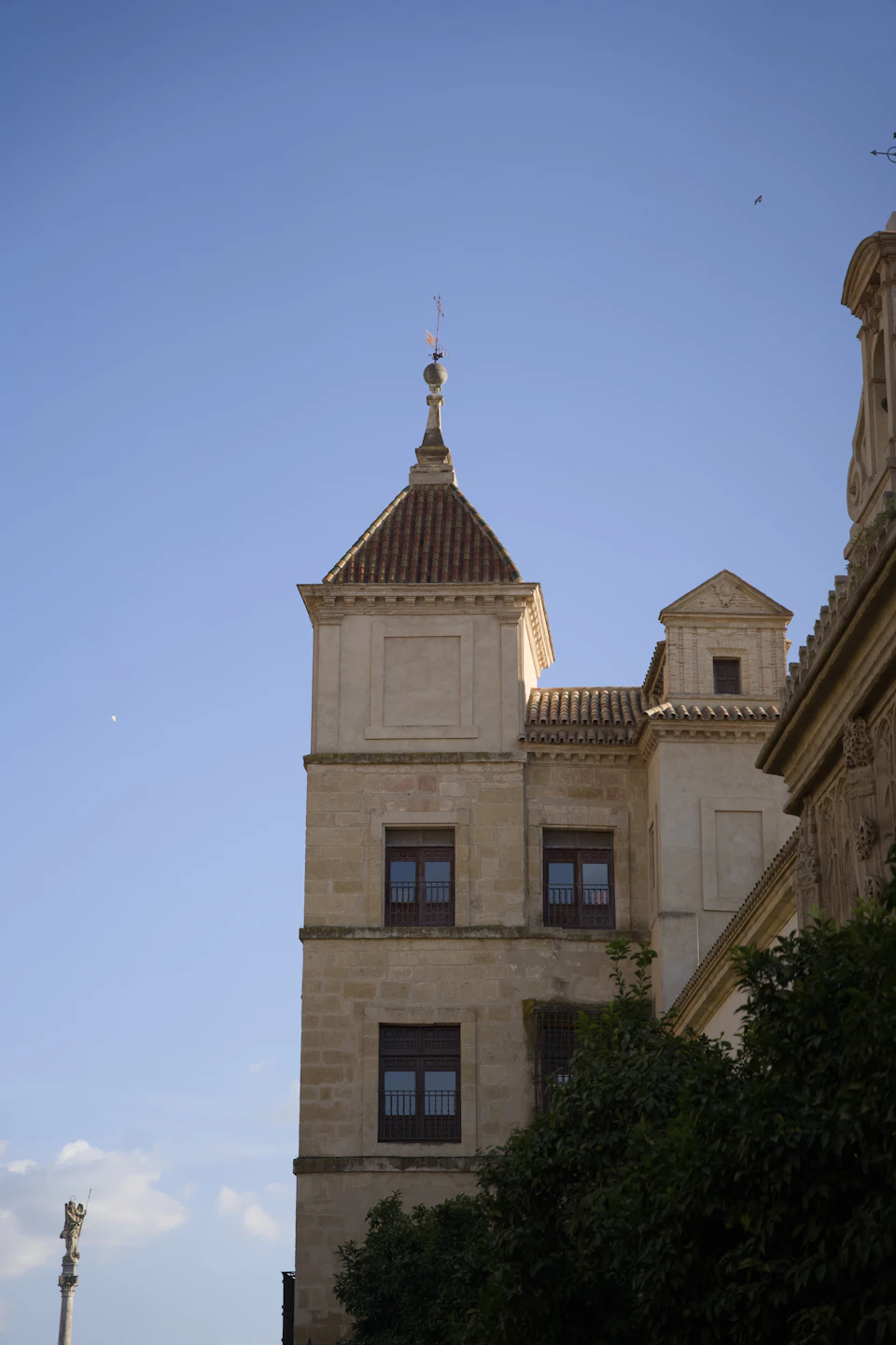 The image depicts a tall, light-colored stone building with a pointed roof and a small tower on top. The building is situated amidst lush green trees, creating a serene atmosphere. A few birds can be seen flying around the structure.