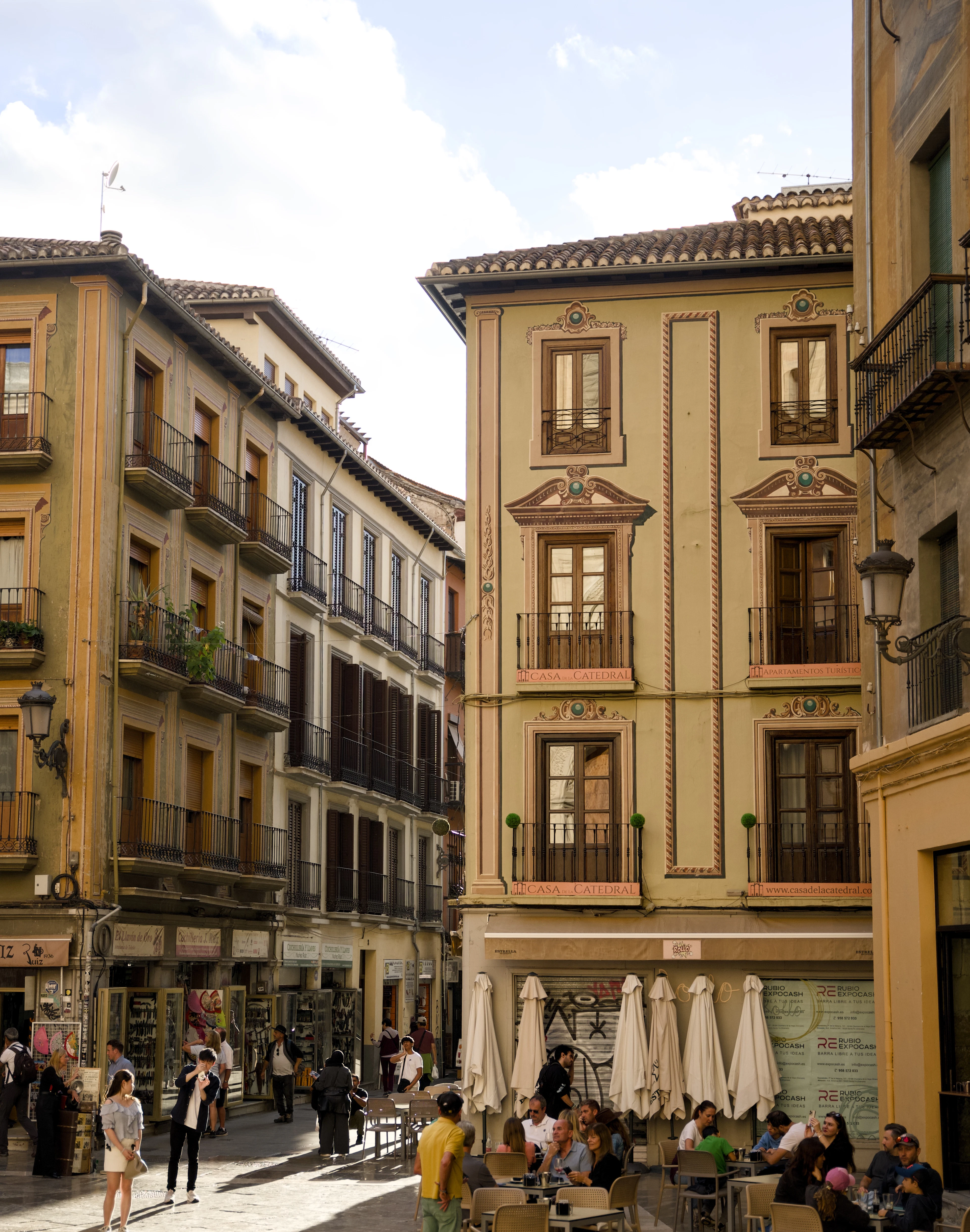Groups eating at cafe tables under white parasols on a sunlit Granada street, ochre townhouses with painted stucco and iron balconies rising above the shopfronts behind them.