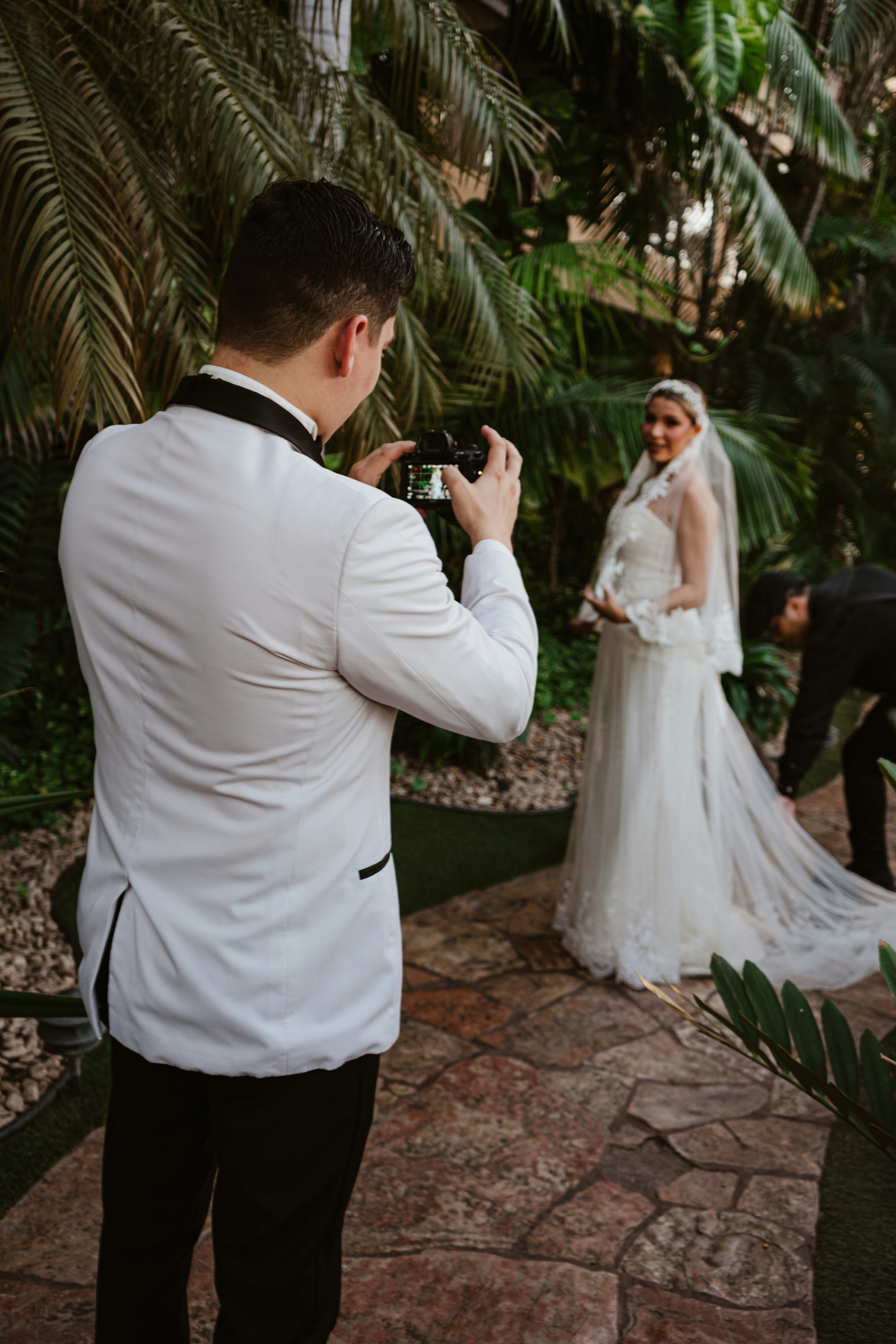 Groom photographs bride in lush garden setting.