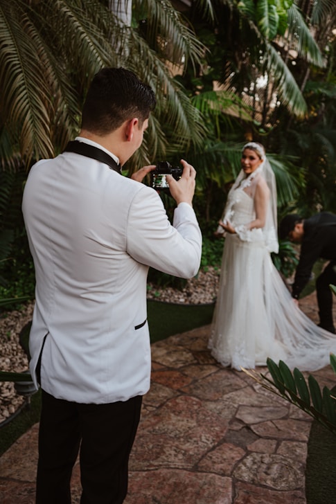 Groom photographs bride in lush garden setting.