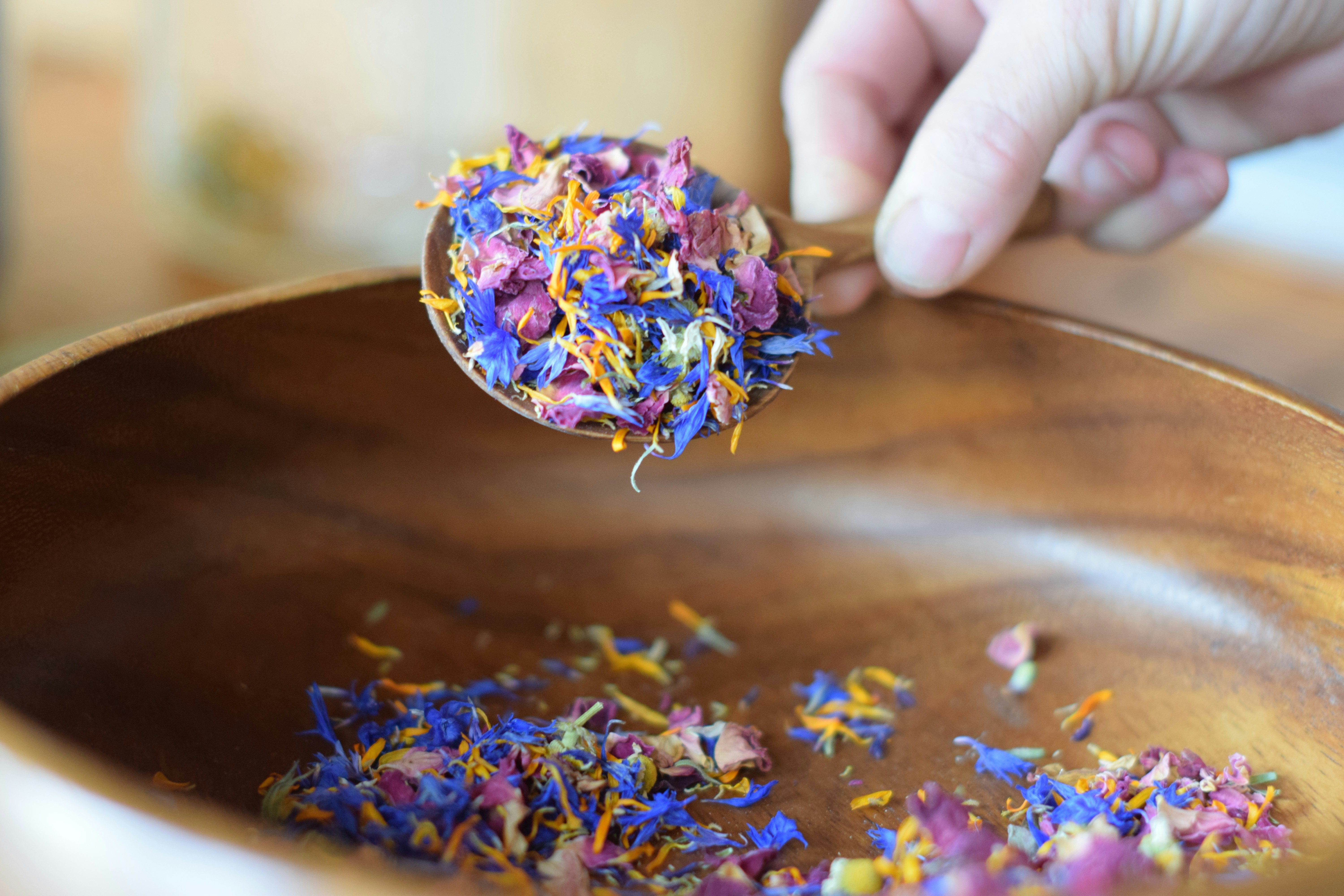 Dried colorful flower petals being scooped into a wooden bowl.