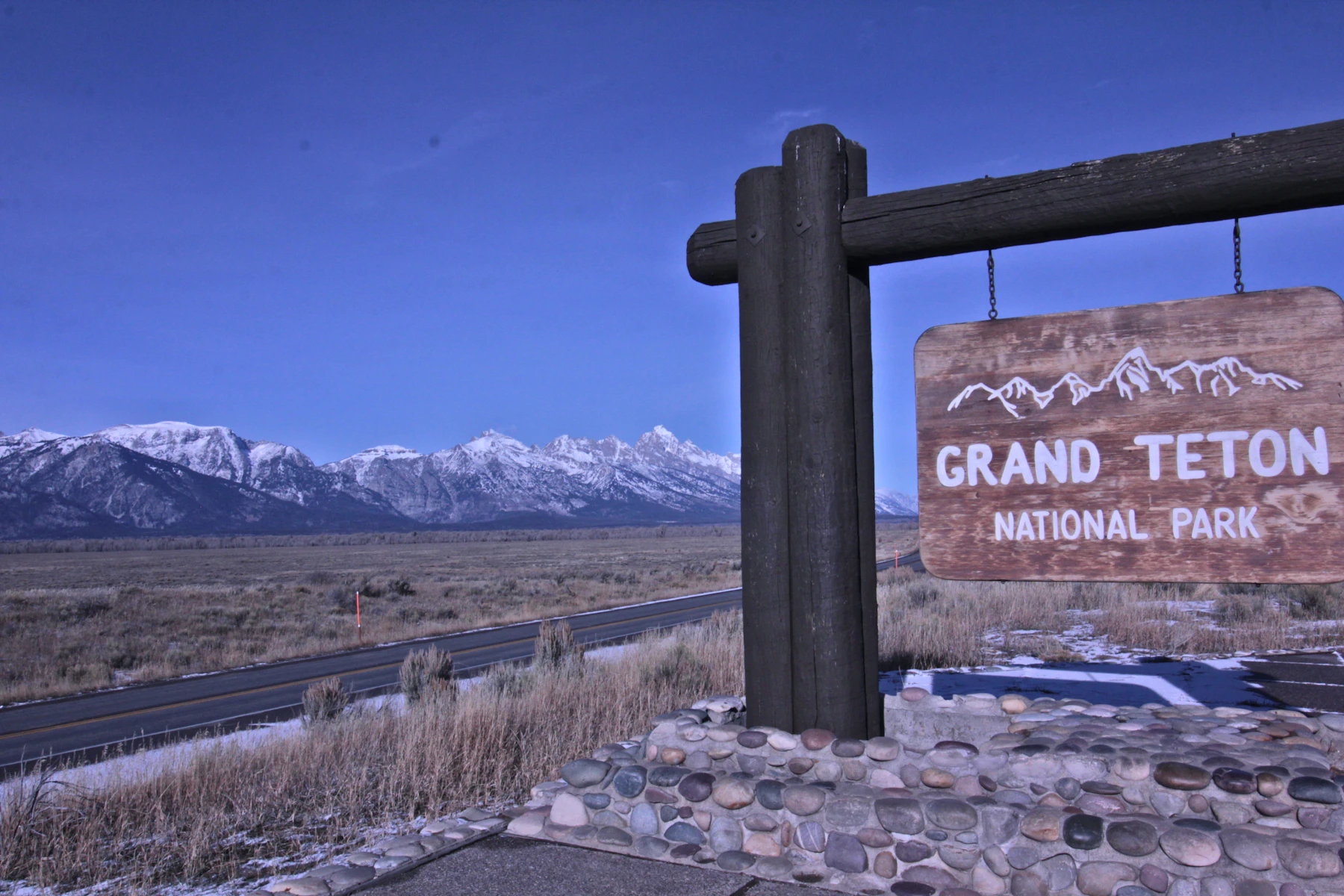 The Grand Teton National Park sign hanging from a log post, with the snow-covered Teton Range behind it