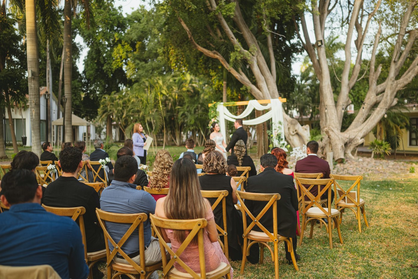 Wedding guests seated on wooden chairs on a lawn under trees watching a ceremony under a flower arch
