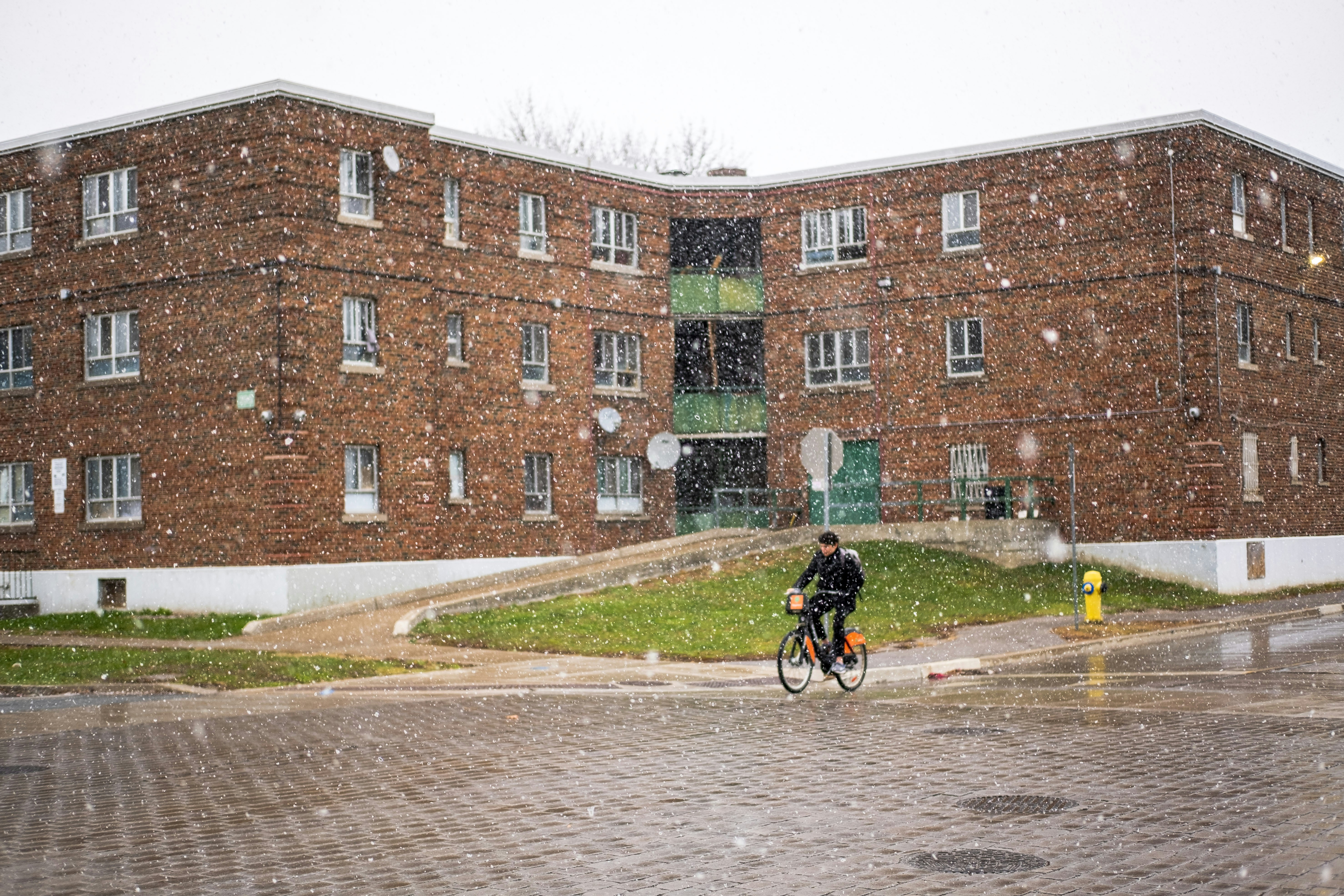 Man rides bicycle in front of brick building during snowfall