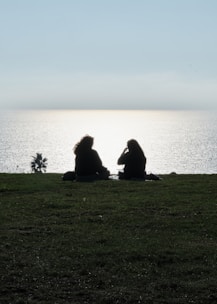 Two people sit on a grassy hill overlooking the ocean.