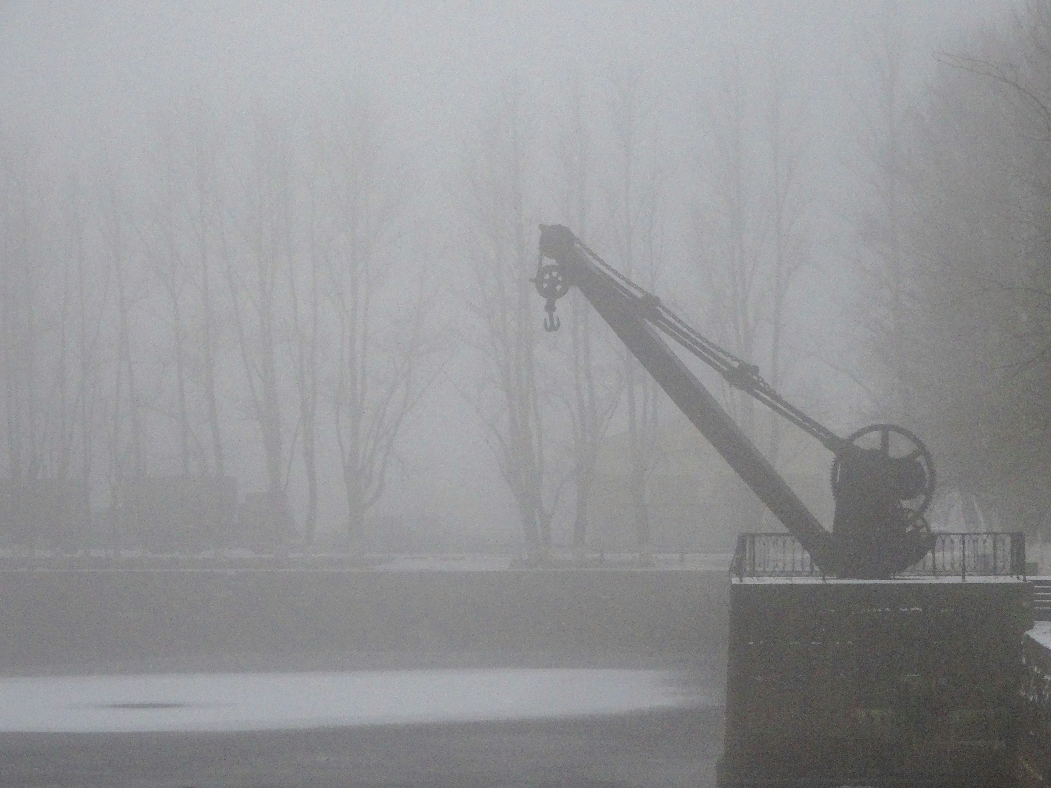 Ein alter Kran steht im dichten Nebel am Wasser.