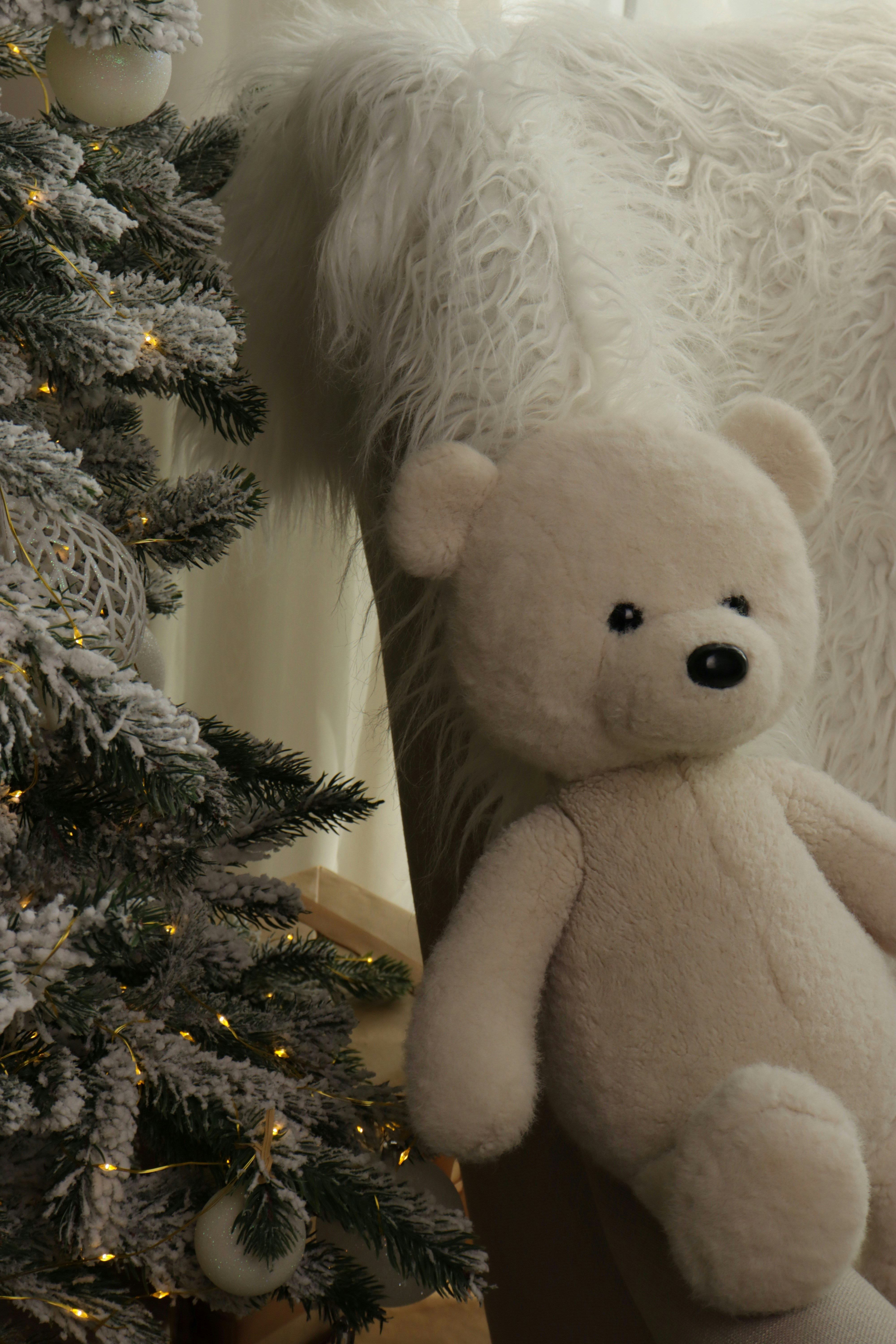 A white teddy bear sits by a decorated christmas tree.