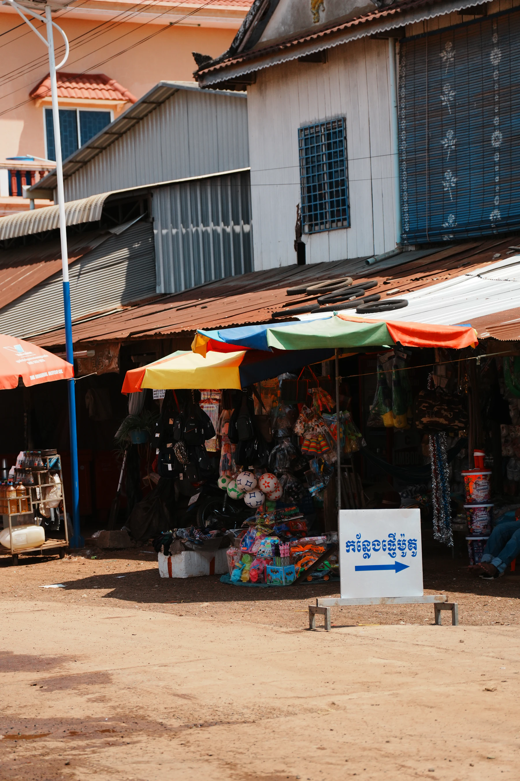 A compact roadside market stall displays colourful goods beneath umbrellas and corrugated roofs.