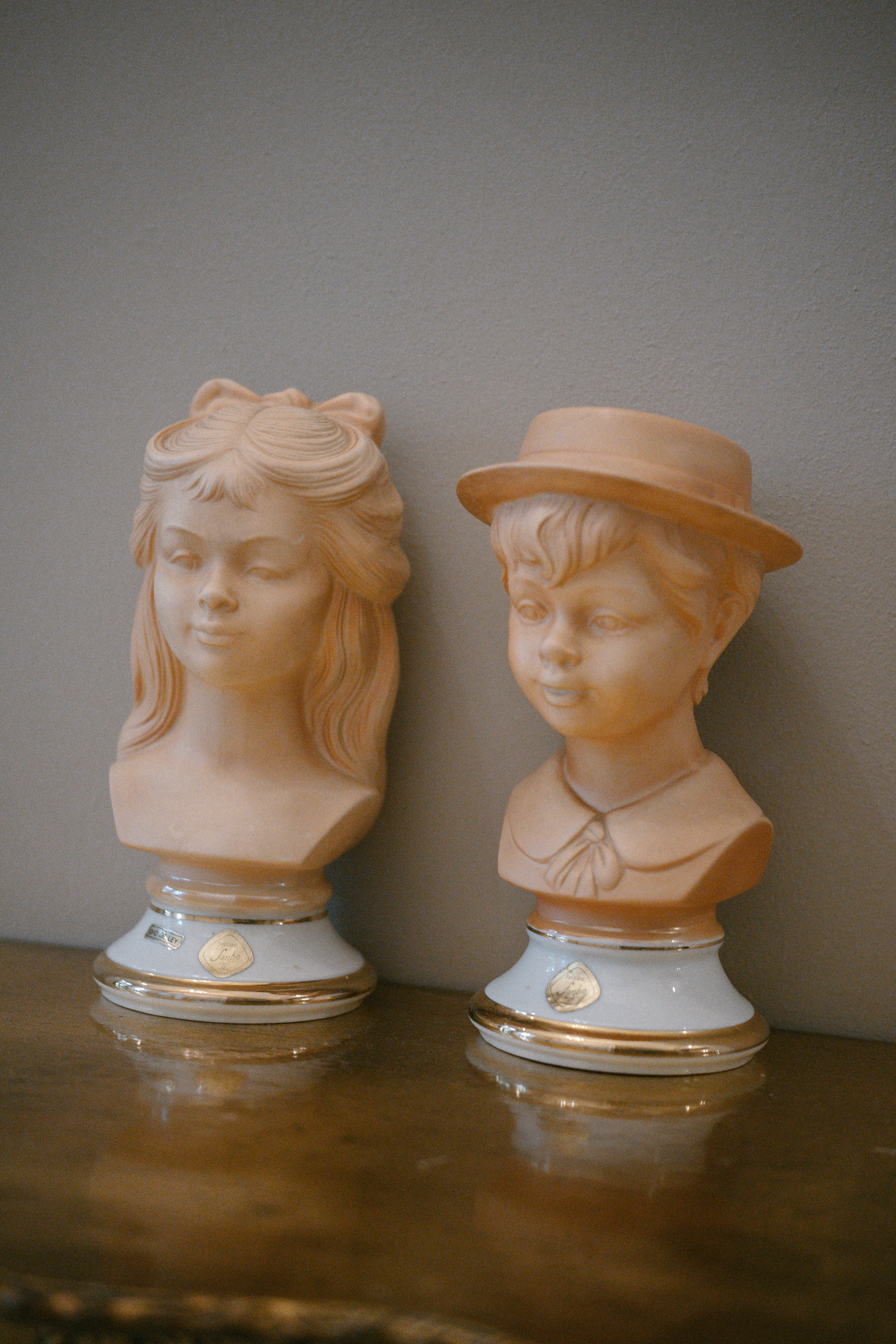 Two unglazed ceramic busts of children, a girl with a bow in her hair and a boy in a brimmed hat, on a shelf