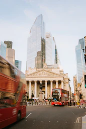 Red buses pass modern buildings in london.