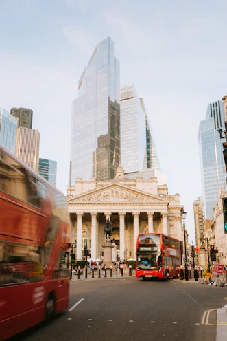 Red buses pass modern buildings in london.