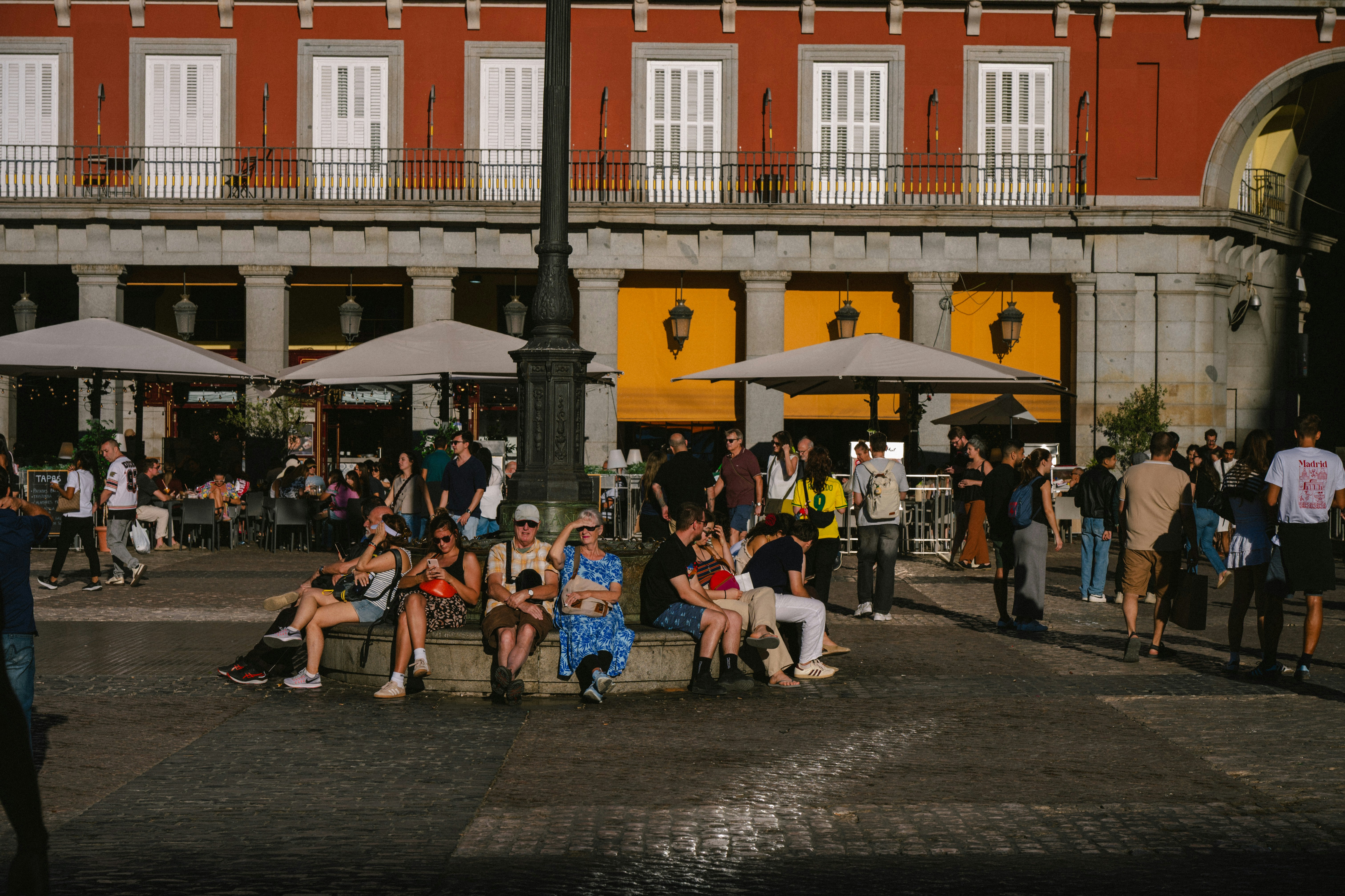 People sitting and socializing in a lively city square with outdoor cafes and a red building in the background.
