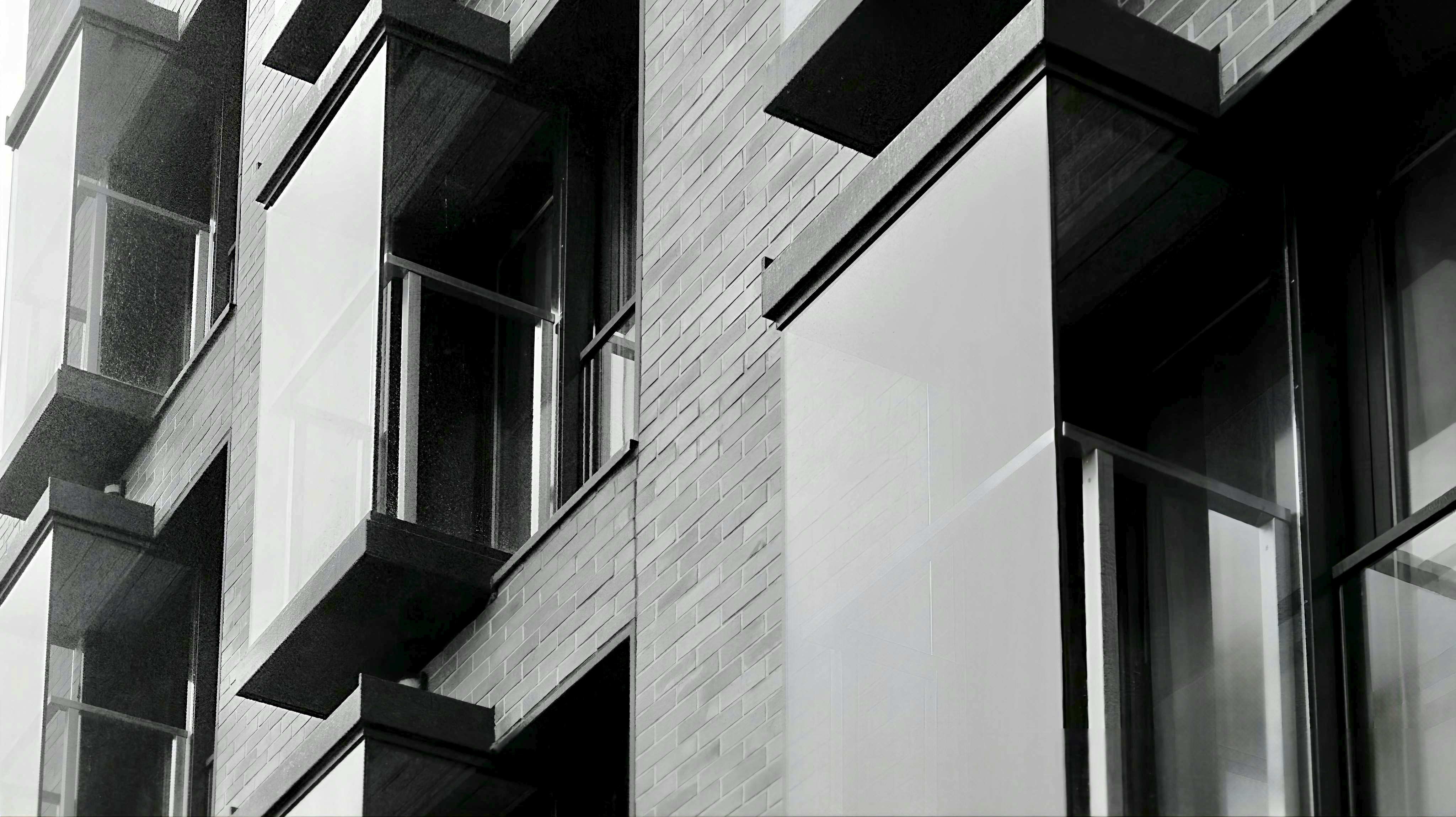 A monochrome, close-up view of a modern building facade featuring textured brick walls, large reflective glass panels, and dark geometric projections.