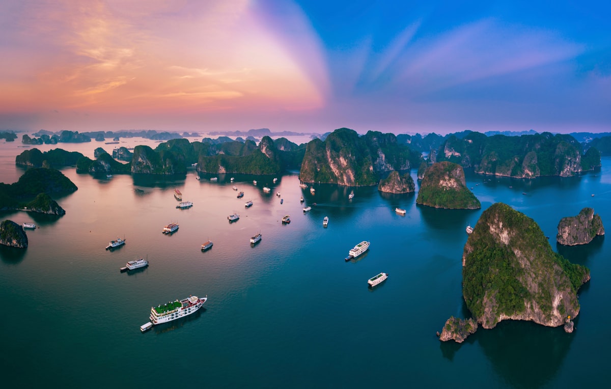 Numerous limestone karst islands rise from emerald green waters at sunset, with the sky painted in shades of orange, violet, and gold.. Photo by Vivu Vietnam on Unsplash.