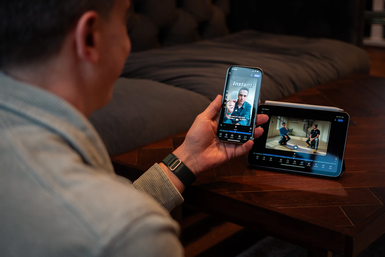 A man on a sofa watching a video on his phone with a tablet beside him