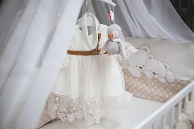 A delicate white baby dress hangs in a crib.