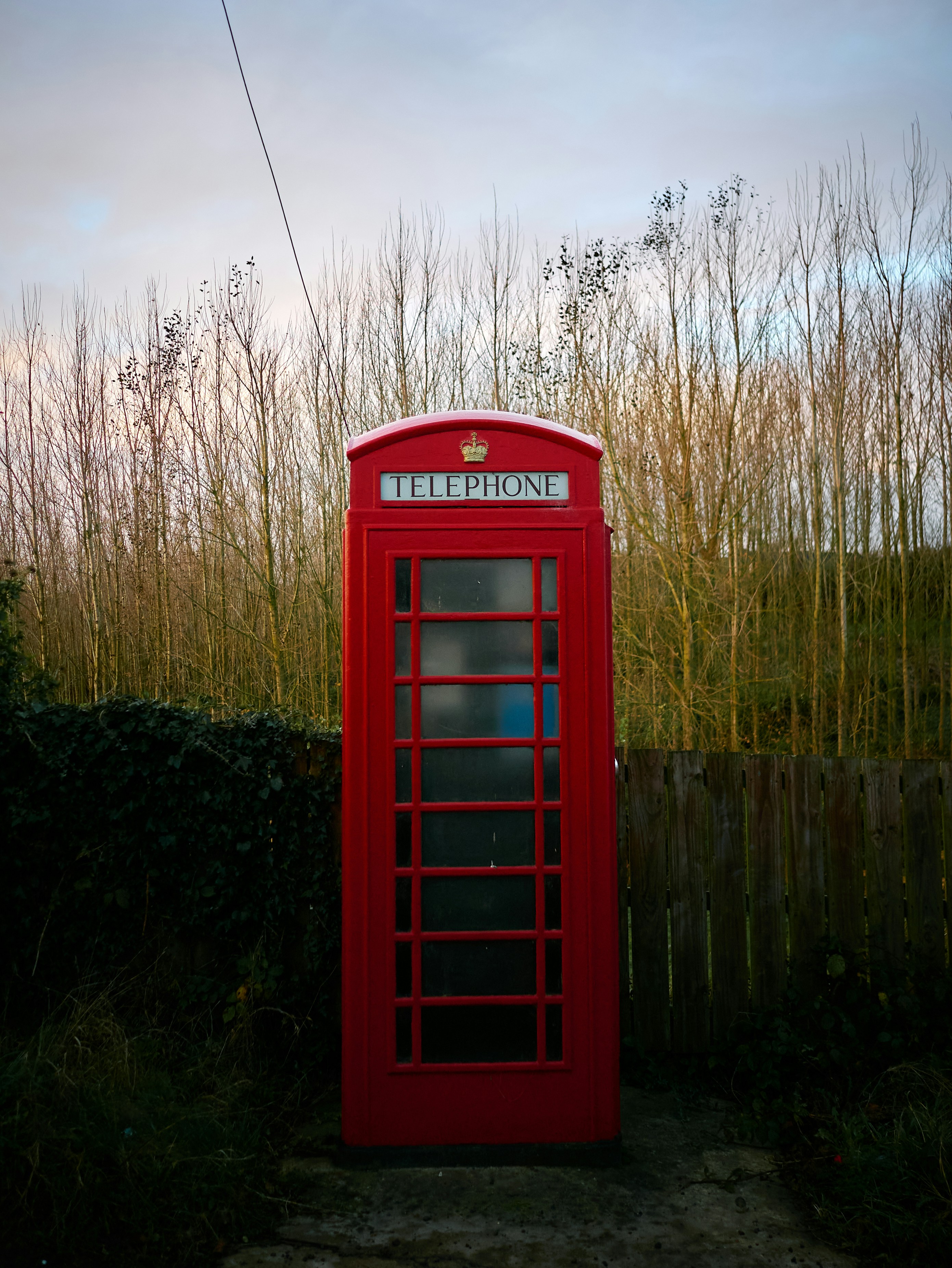 A red telephone booth stands outdoors with trees behind it.