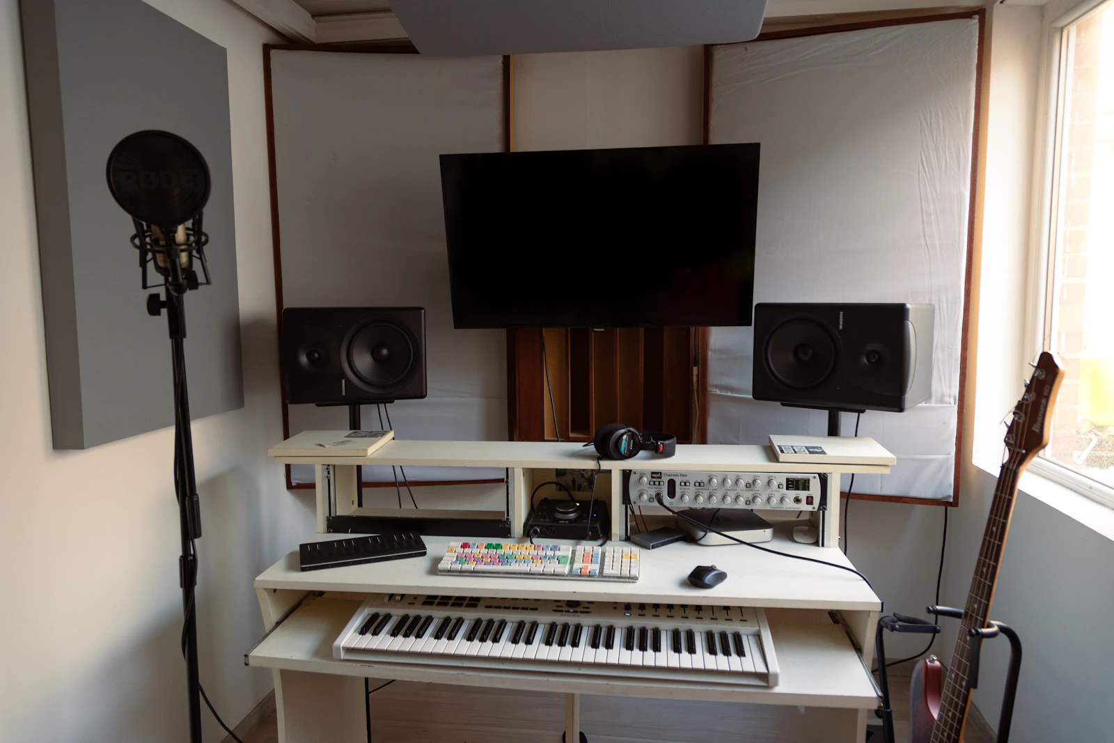 A bright home recording studio with a keyboard, studio monitors, a microphone on a stand and a guitar