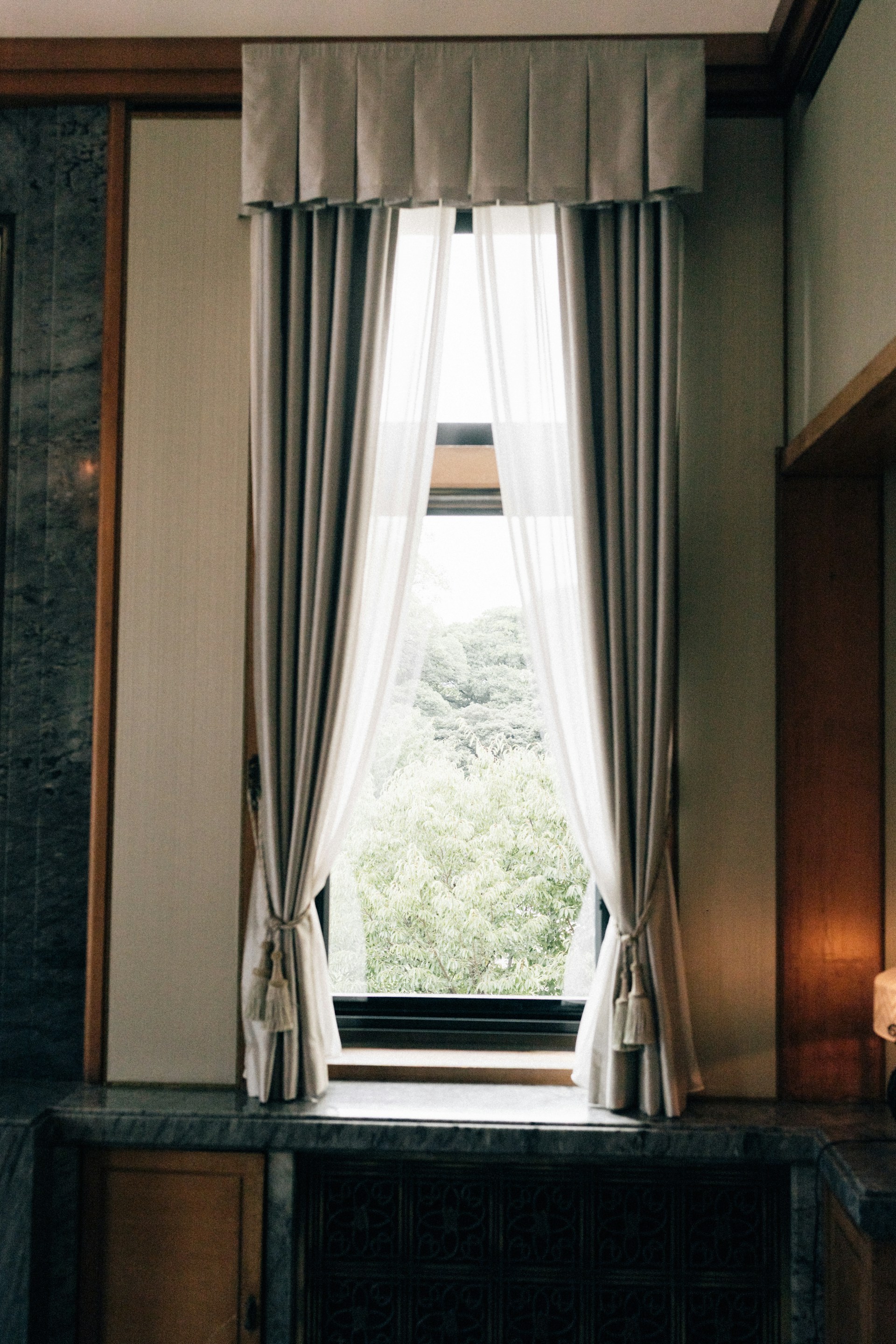 Tall window with light grey curtains and view of trees.