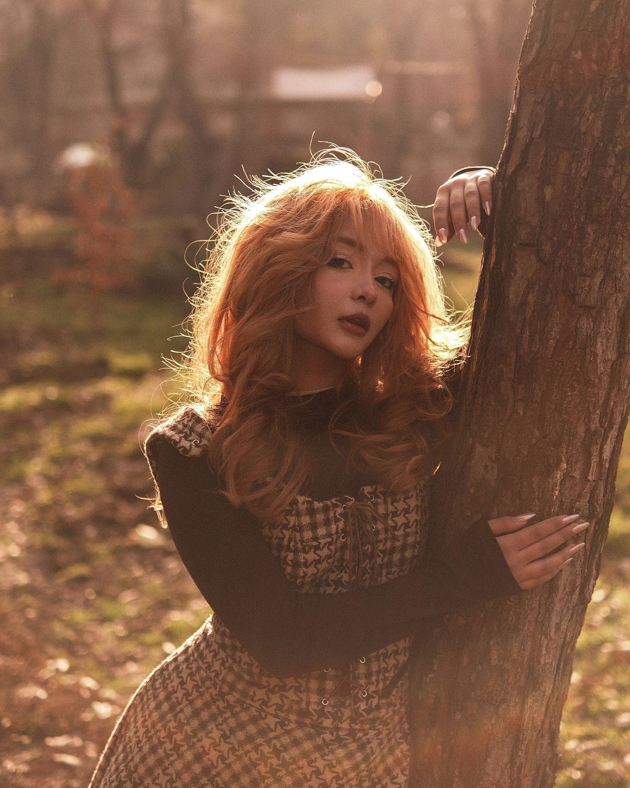 Woman with red hair leans against a tree outdoors.