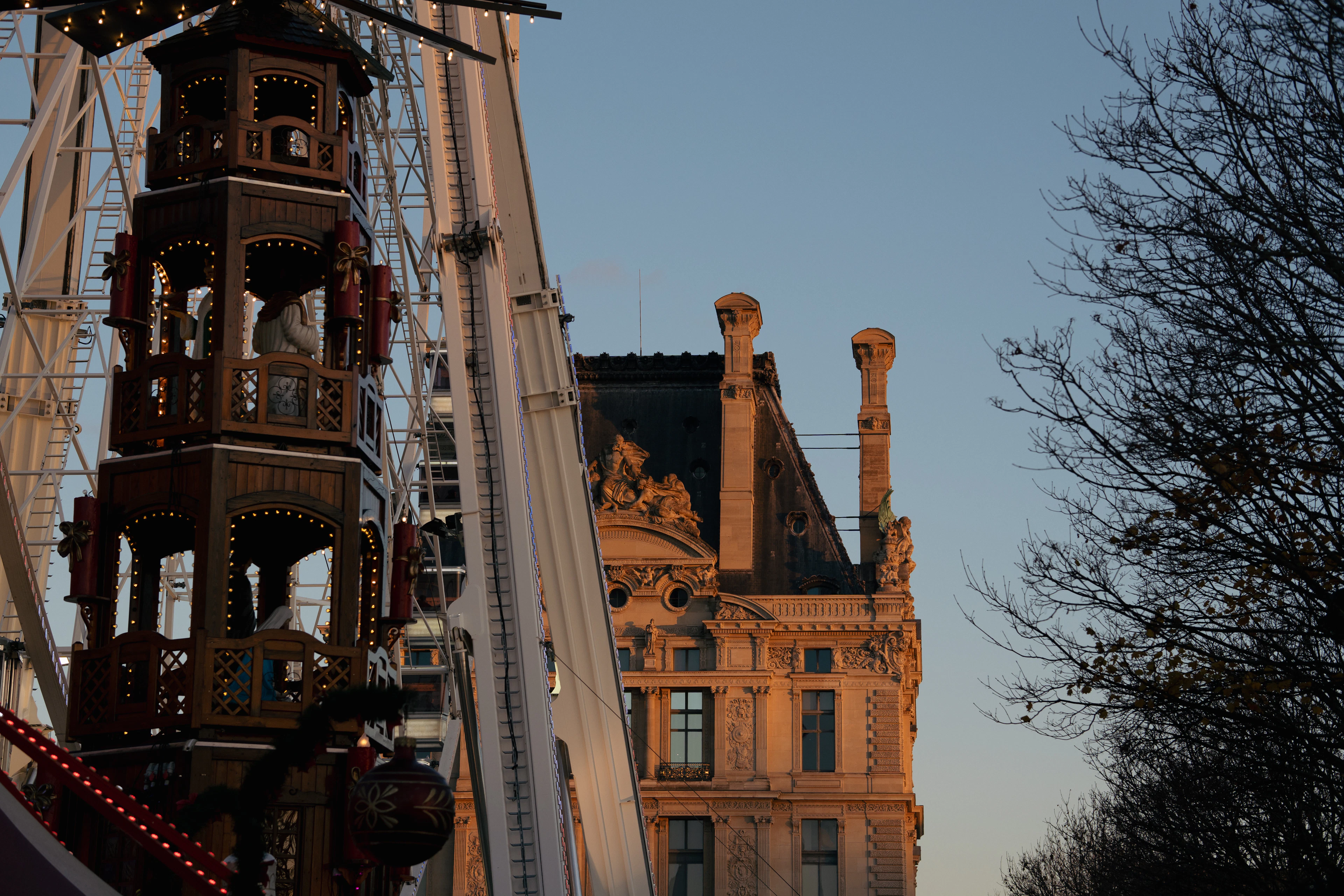 A lit wooden Christmas pyramid and Ferris wheel at the Tuileries winter fair, with a Louvre pavilion glowing at dusk
