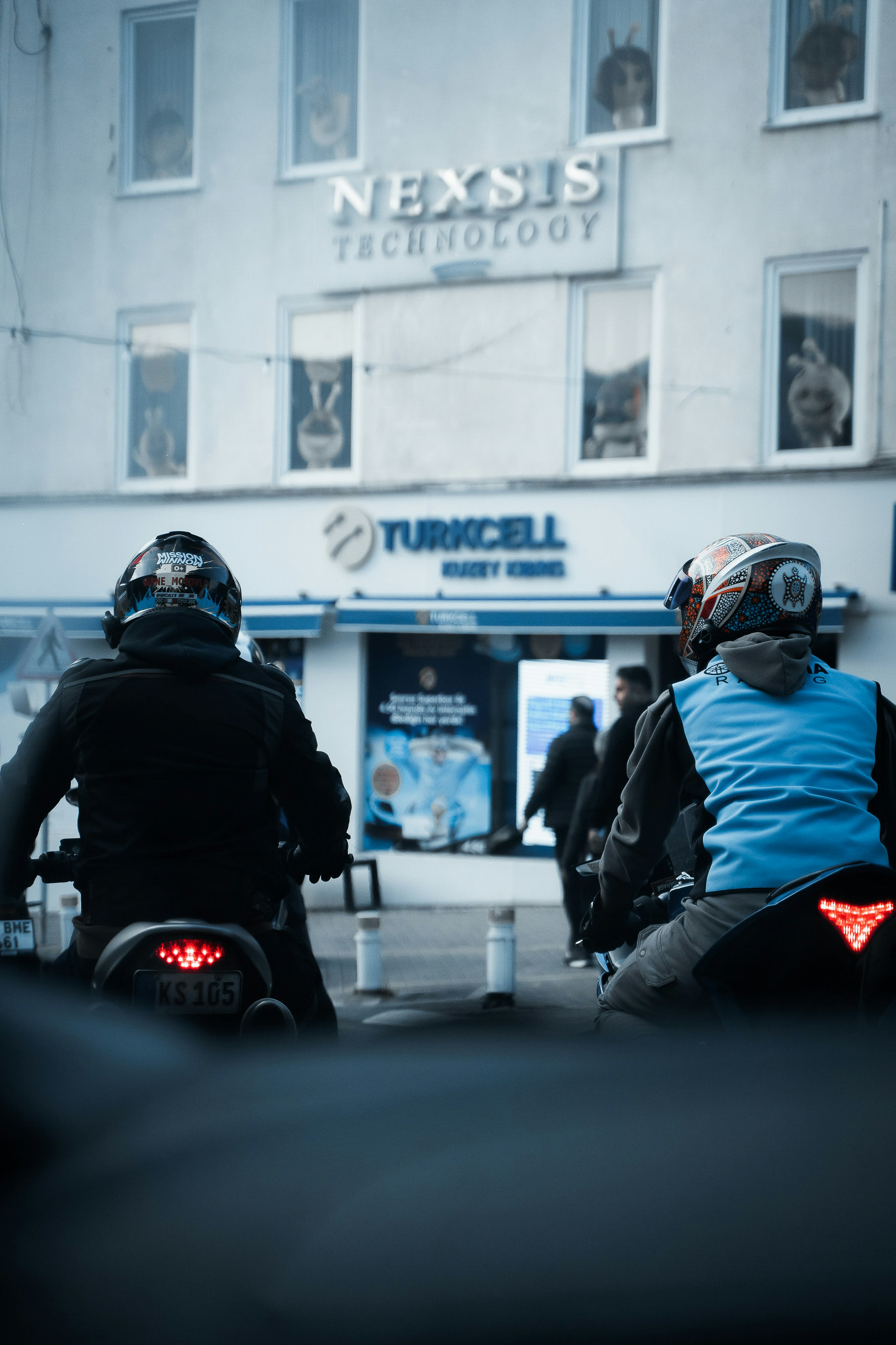 Two motorcyclists wait outside a building.