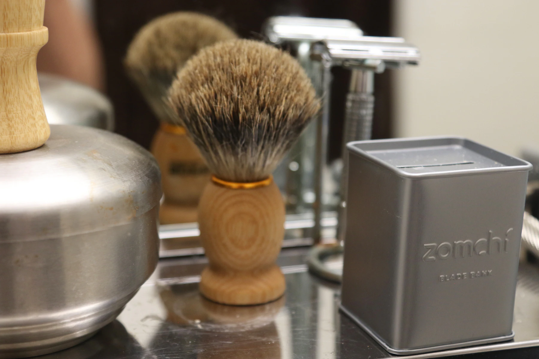 A badger brush with a wooden handle beside a safety razor on a stand and a steel blade tin, in front of a mirror