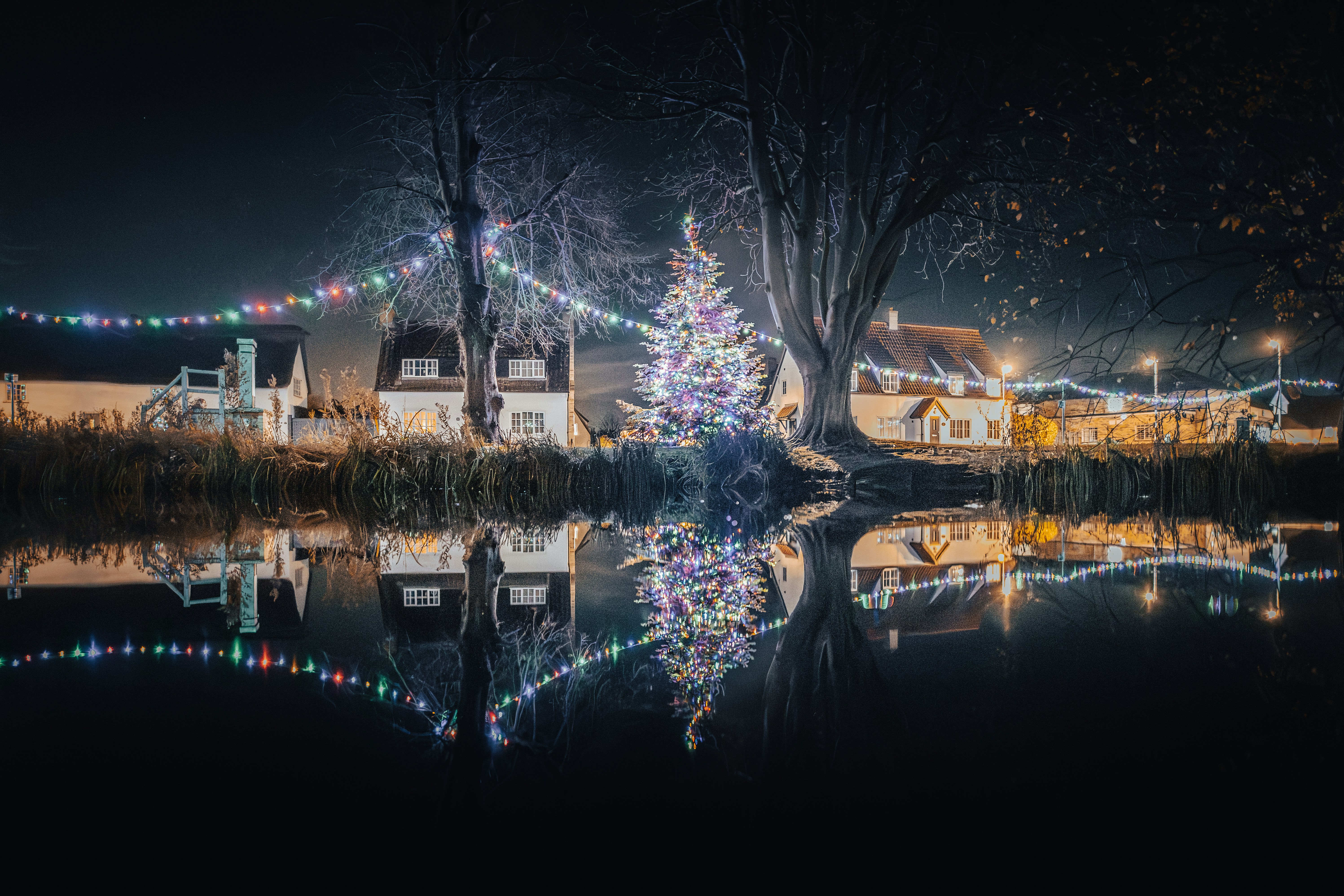 Dekorierte Weihnachtsbäume und Häuser spiegeln sich nachts im Wasser.