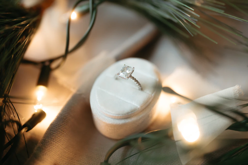 An engagement ring in a cream velvet box surrounded by pine branches and warm string lights