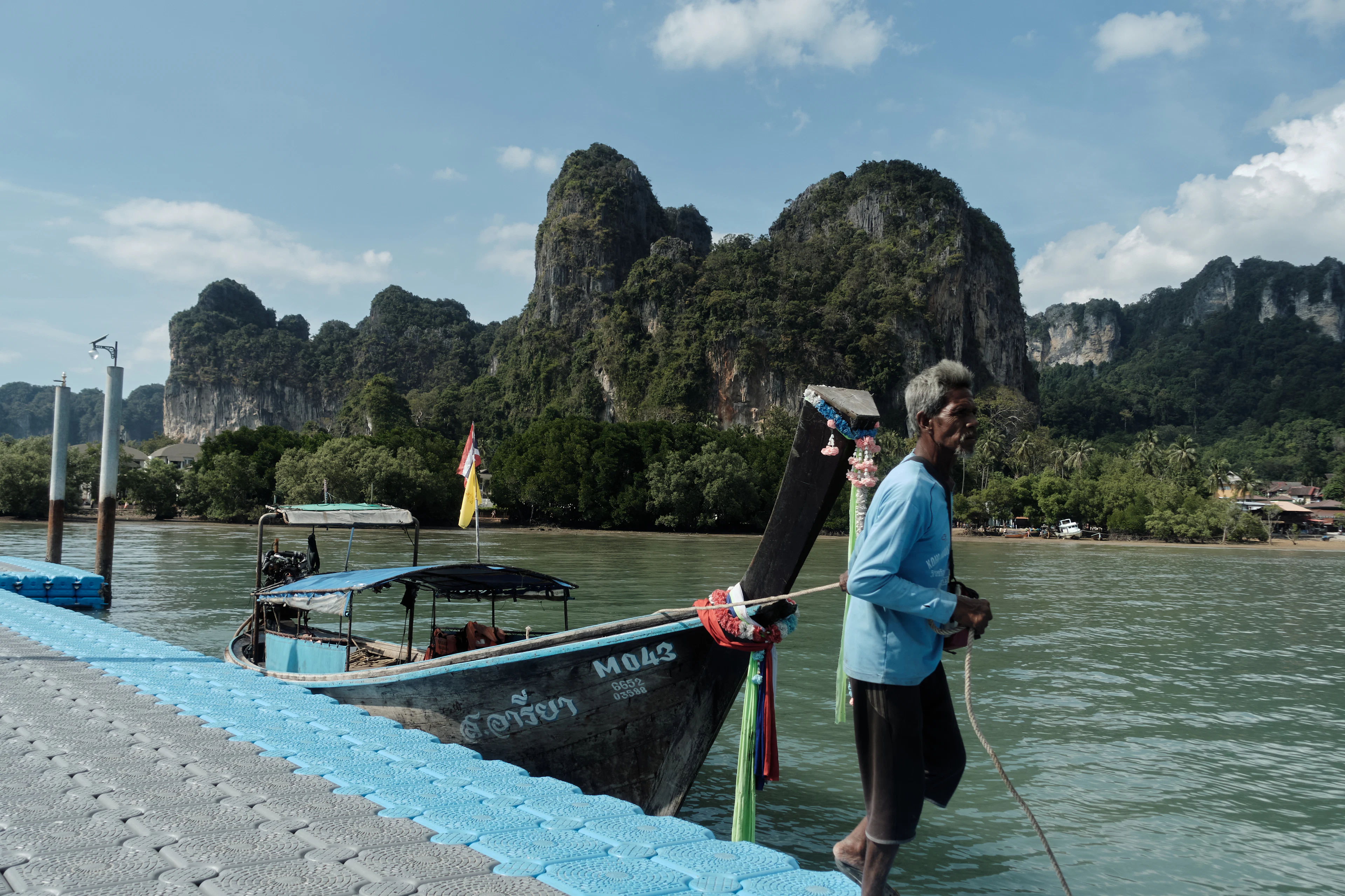A boatman rigging his longtail boat at a pier in Krabi, mangroves and jagged karst mountains rising across the water.