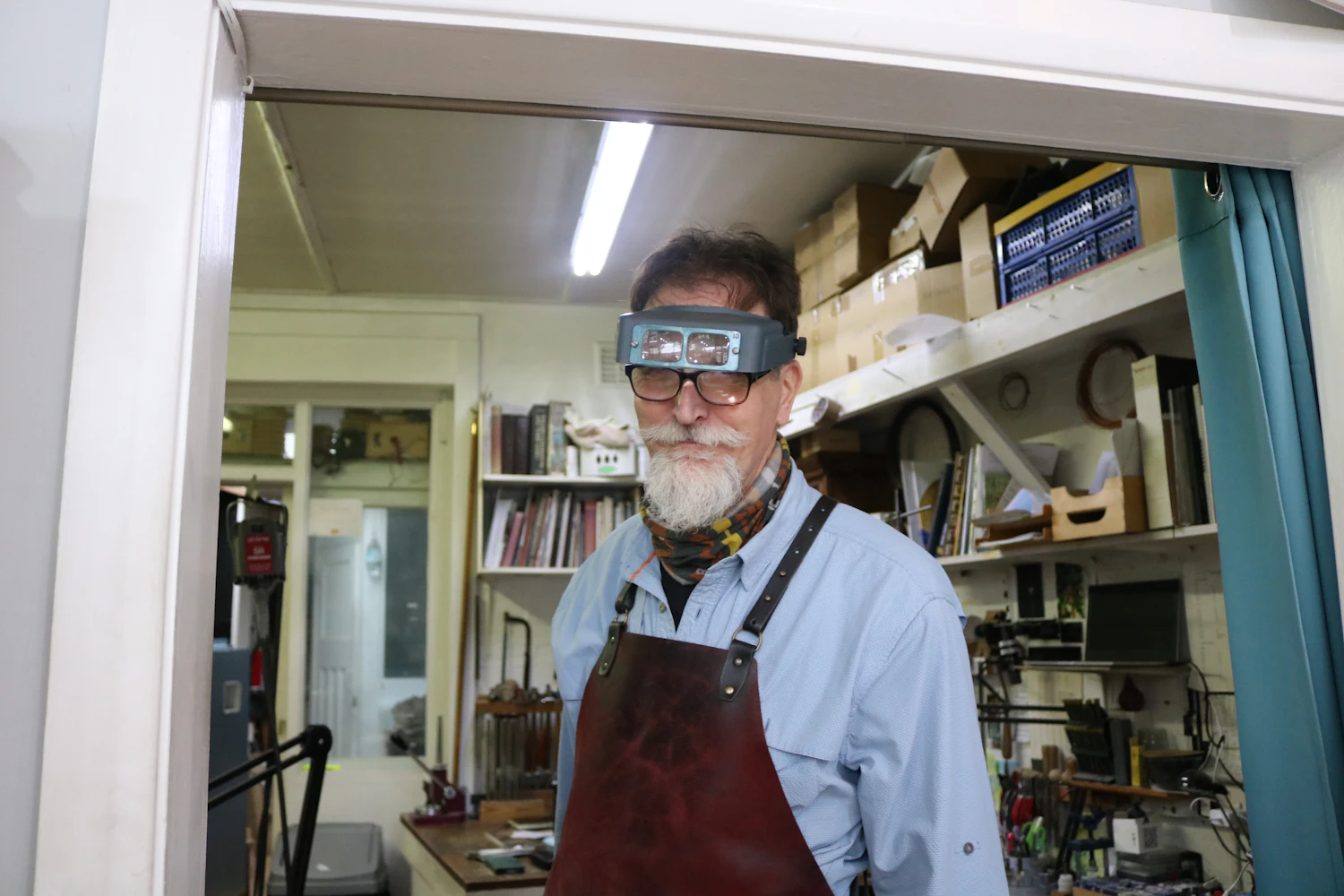 A watchmaker in an apron and magnifying glasses standing in a workshop doorway