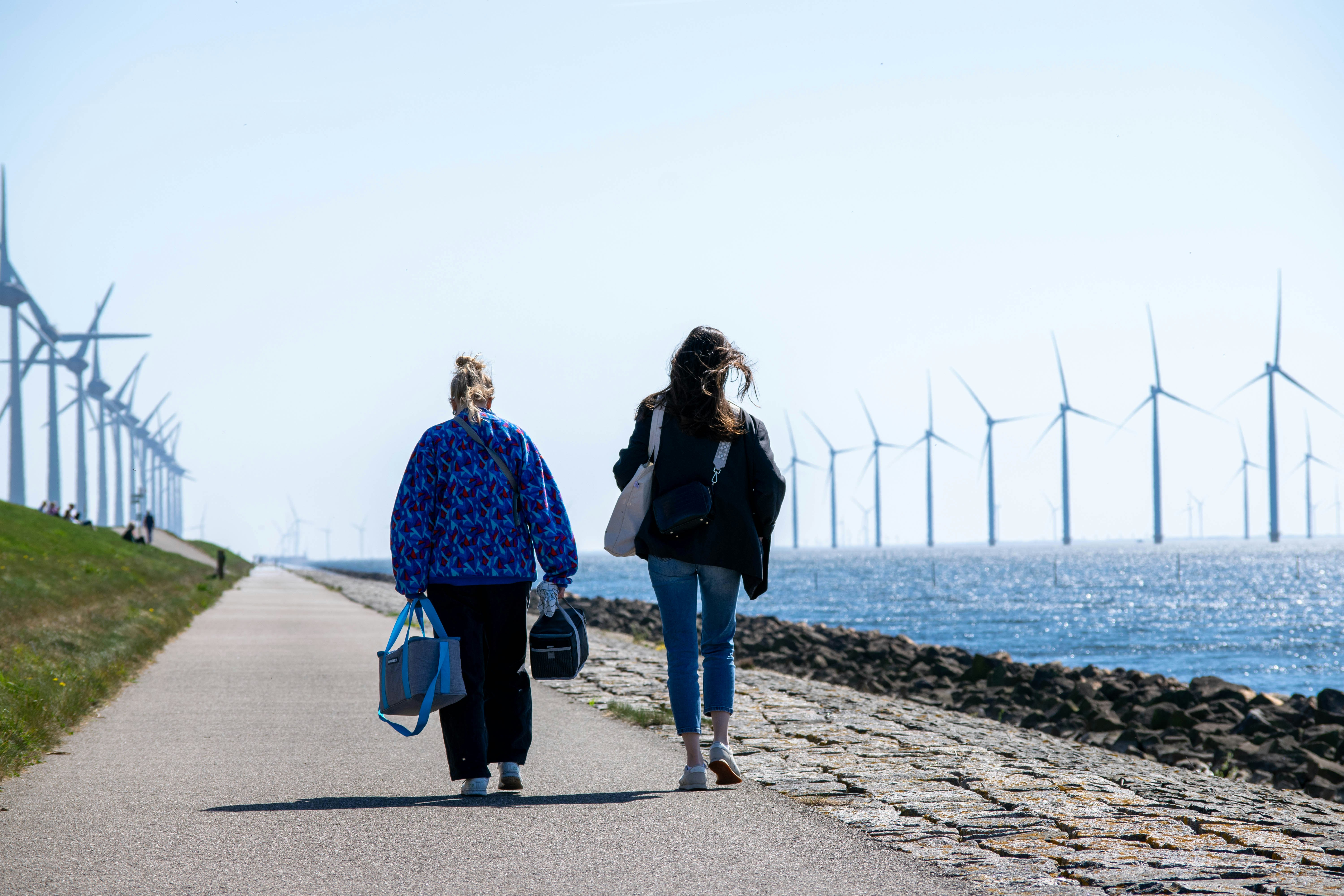 Two women walk along a path with wind turbines offshore.