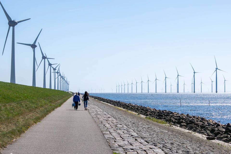 Two people walk along a coastal path lined with wind turbines near the ocean