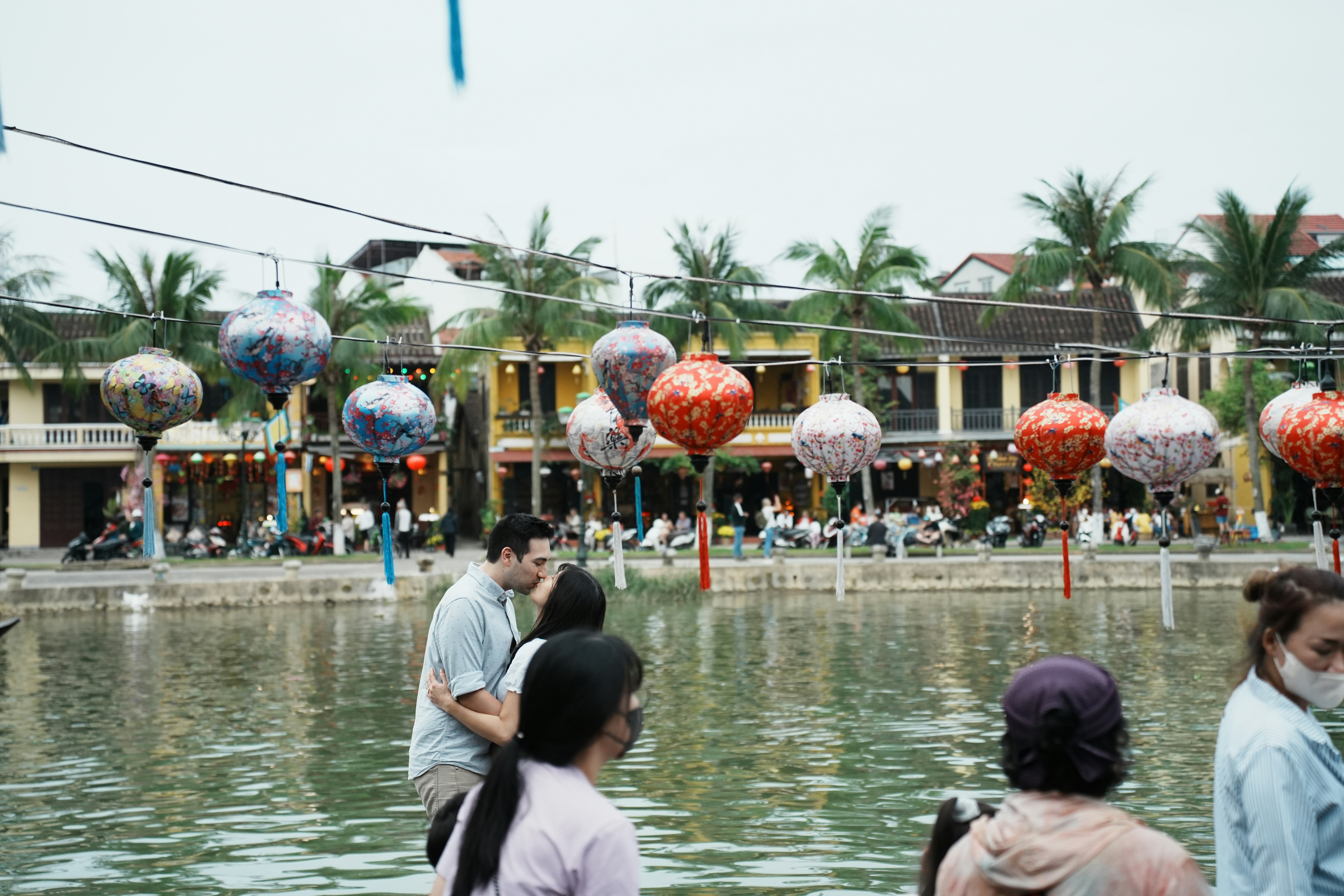People by river with colorful lanterns and buildings.