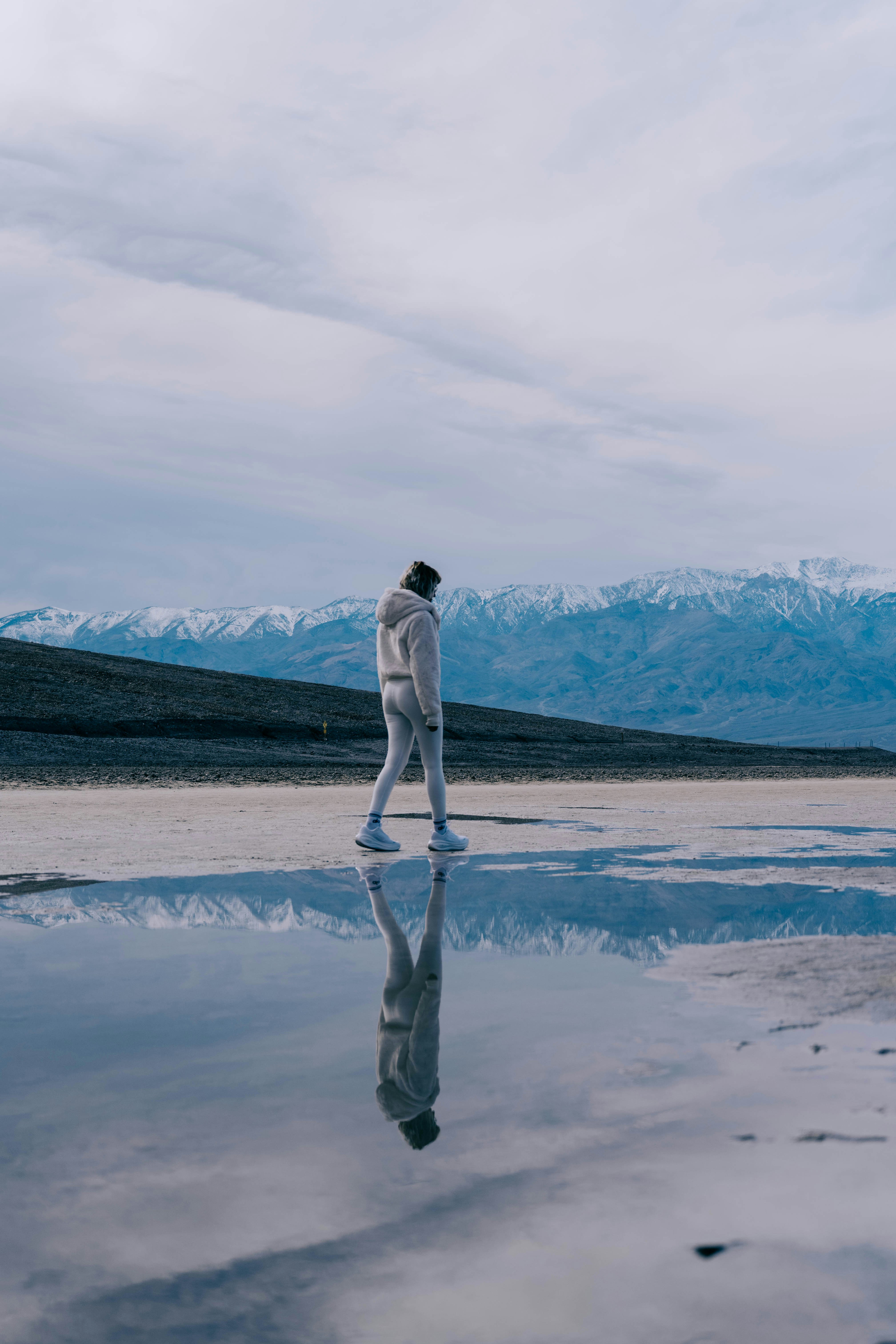 Person walking by a reflective body of water with mountains.