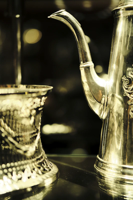 Close-up of an ornate silver coffee pot and cup, showing the polished surface and gauge of the metal