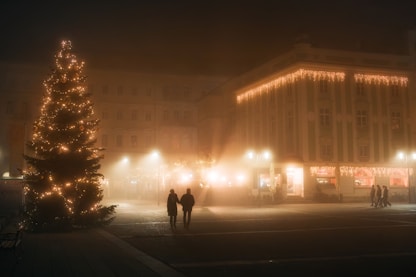Misty Christmas. Time to walk together — But why not on a global scale? (A moody, foggy night scene in a town square in Gmunden, Austria during Christmas. A large illuminated Christmas tree glows on the left, while a silhouette of a couple holding hands walks through the mist toward warm city lights.)