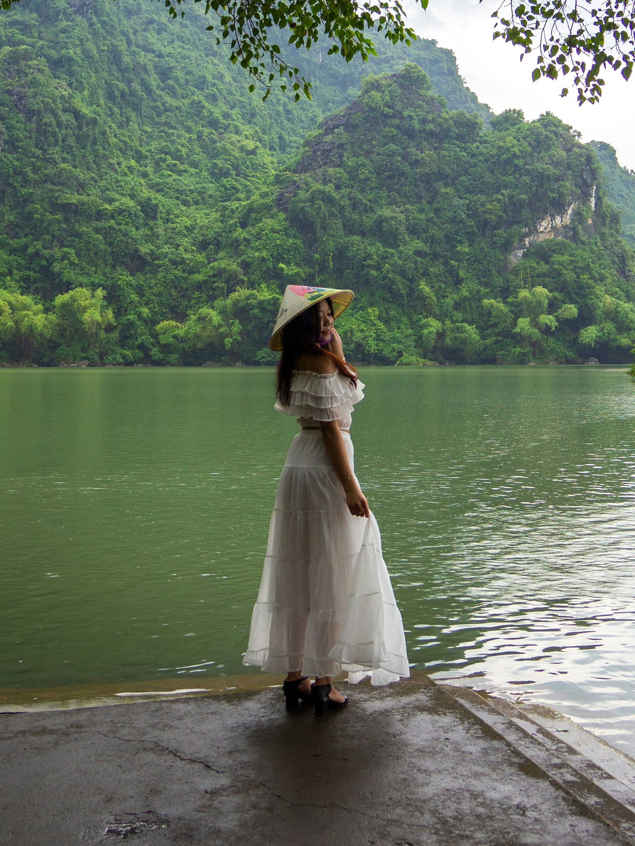 Woman in white dress and conical hat by river.