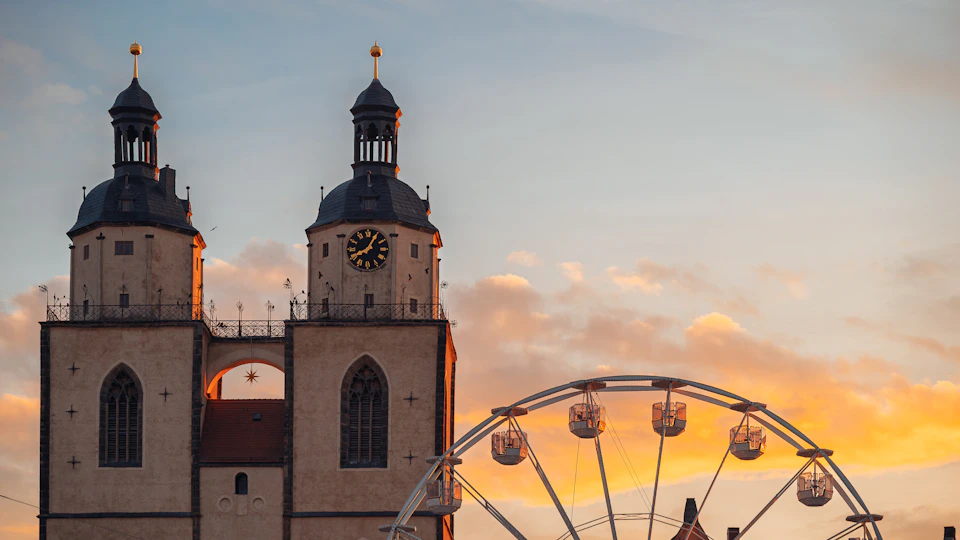The image depicts a large, white church with two tall towers and a clock face on each tower. The church is situated next to a ferris wheel, which is painted in shades of orange and yellow and has multiple circular gondolas attached to it. The sky above the scene is filled with clouds, creating a dramatic backdrop for the architectural elements below.