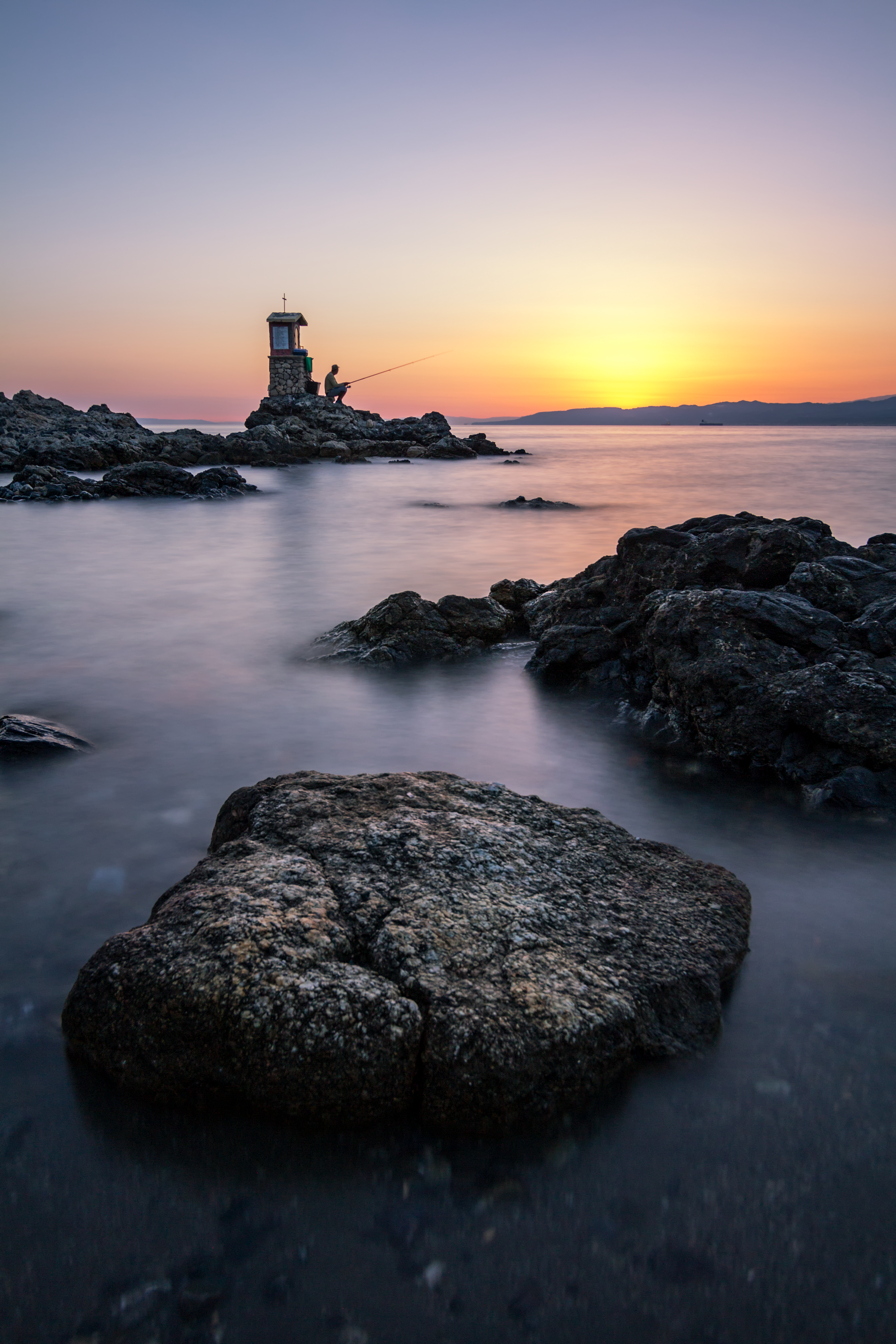 Two people fishing by a lighthouse at sunset.