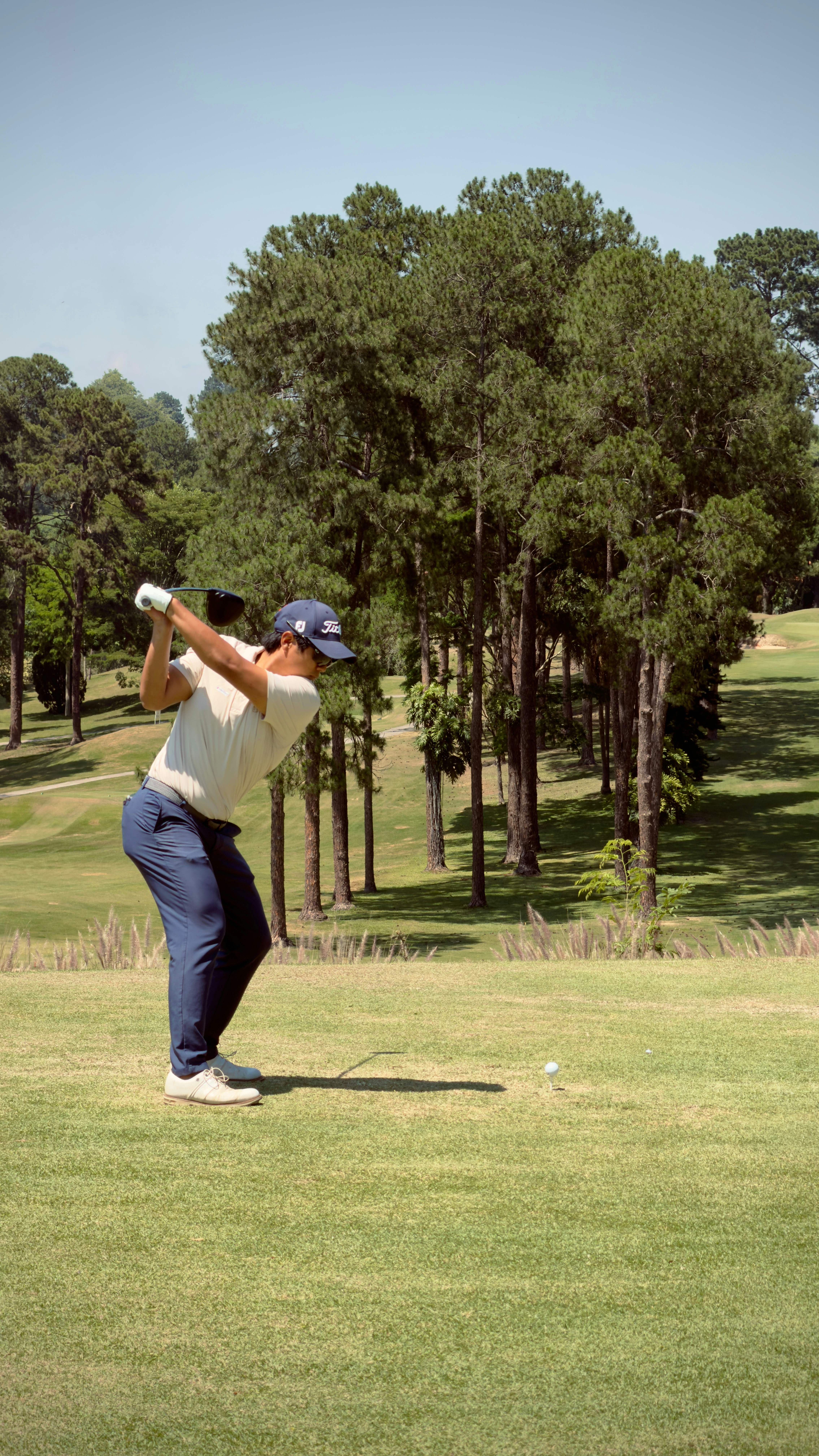 Golfer swings club on sunny day with trees.
